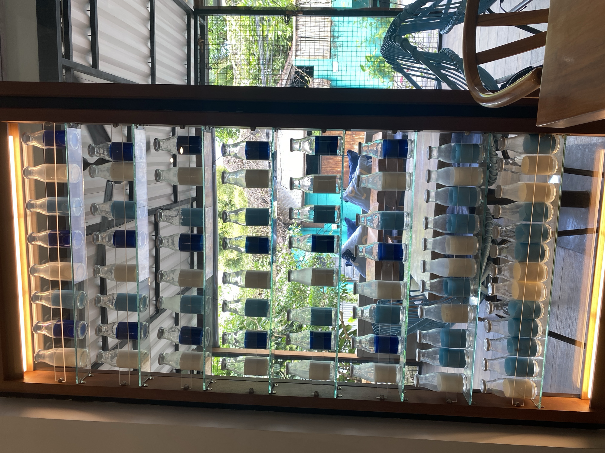 The image shows a decorative window made of glass panels and glass milk bottles filled with blue and white liquid. The window is framed with a dark wood and has a light strip along the left side. The bottles are arranged in a staggered pattern. Through the window, there is a view of a garden and a blue house. The image was taken in North Kuta, Indonesia. In the background, is a chair.