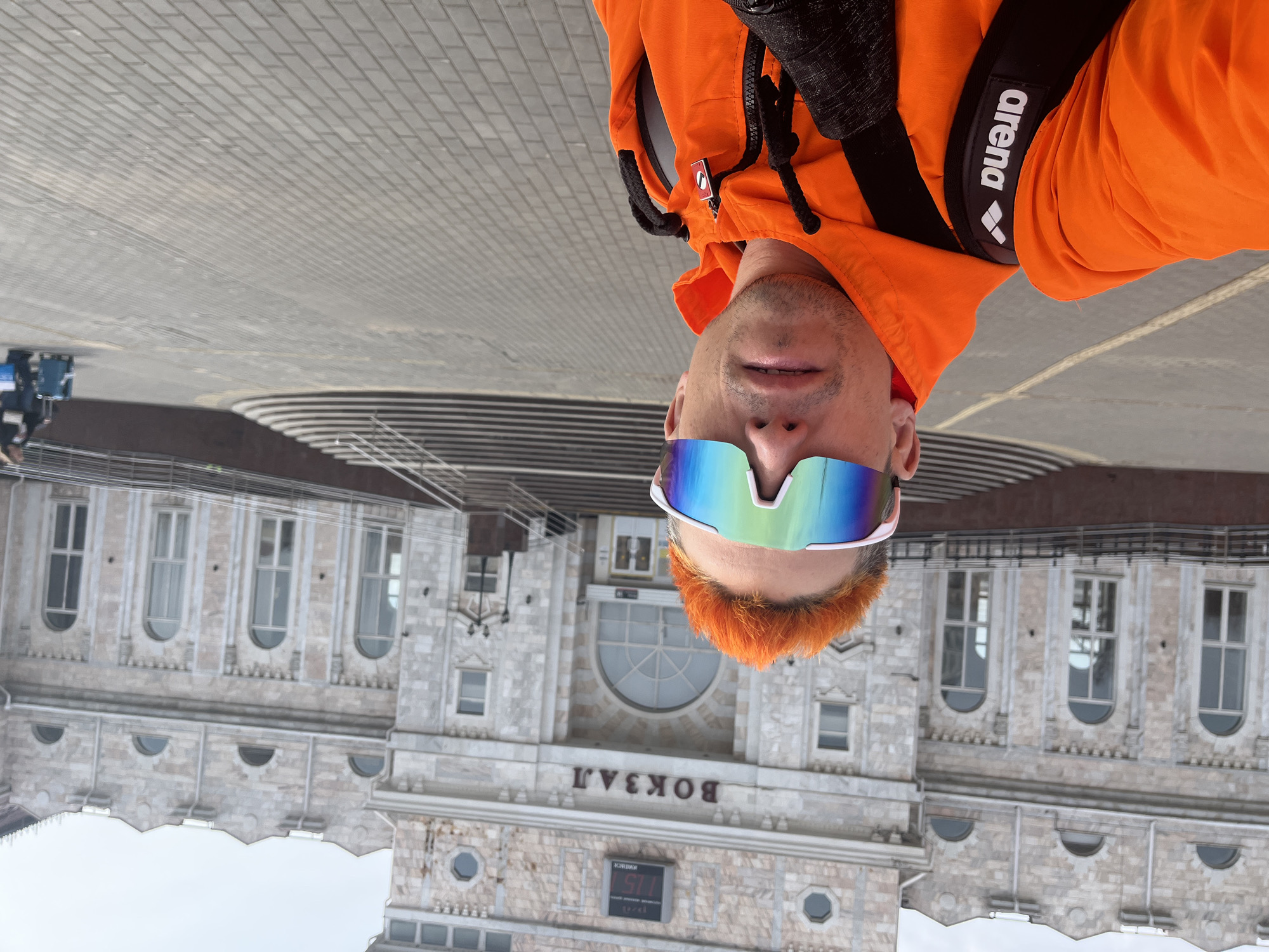 A person in an orange hoodie and futuristic sunglasses stands in front of the Izhevsk railway station in Russia. The building is made of light-colored stone with symmetrical windows and architectural details. Above the entrance is the word "ВОКЗАЛ" in Cyrillic. A digital clock displays the time "5:11". The person is wearing a black backpack with the word "arena" printed on it. The perspective suggests the photo was taken from a low angle and is now upside down. The ground is paved with gray bricks. There is a building to the left.