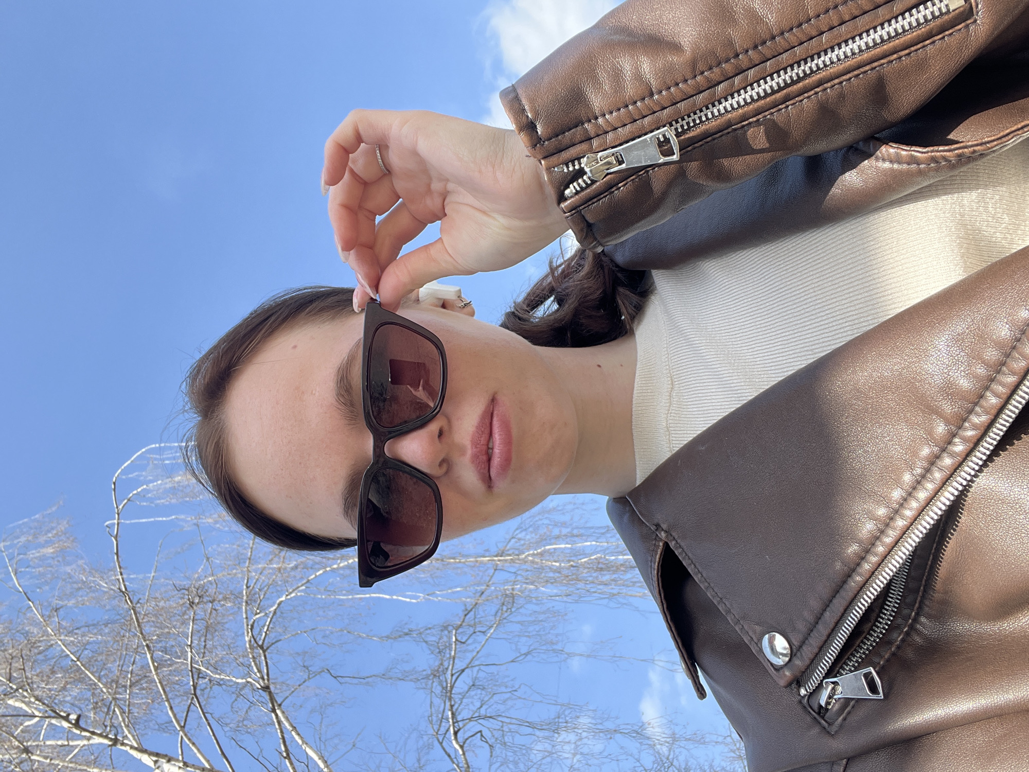 A young woman is posing in an upward-angled shot outdoors in Chaikovsky, Russia. She is wearing a brown leather jacket and sunglasses. The background shows a clear blue sky with a few scattered clouds, and bare tree branches. The woman's hand is touching the frame of her sunglasses.