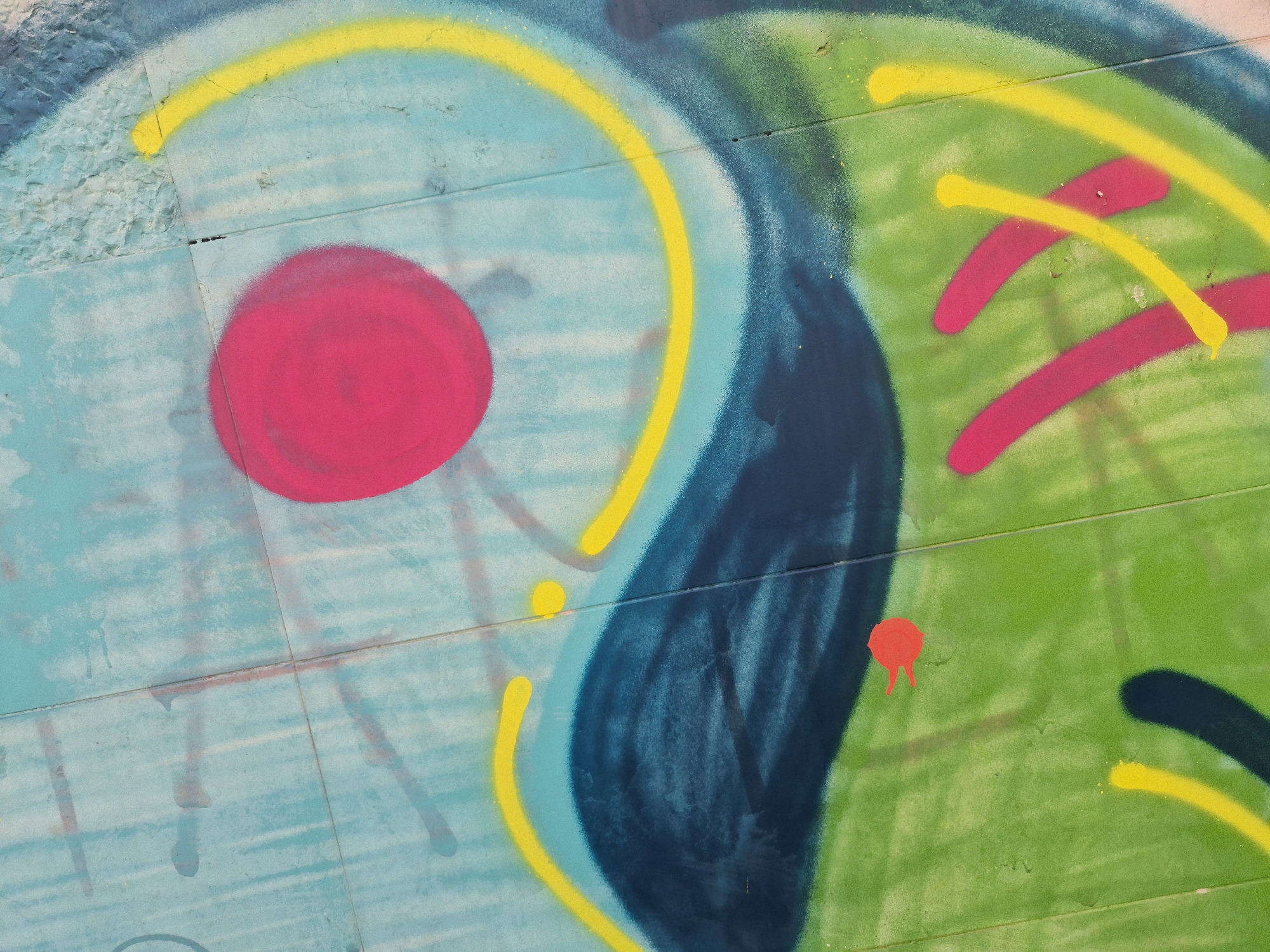 A close-up, abstract view of graffiti art on a textured wall in Taranto, Italy. The graffiti features bold, overlapping spray-painted shapes in vibrant colors, including a large pink circle, arcs of yellow and magenta, and a prominent dark blue curve against a background of light blue and green. The wall surface appears to be made of tiled panels with visible grout lines and some signs of wear and tear. The overall impression is of an urban art installation, with no people or specific actions depicted.