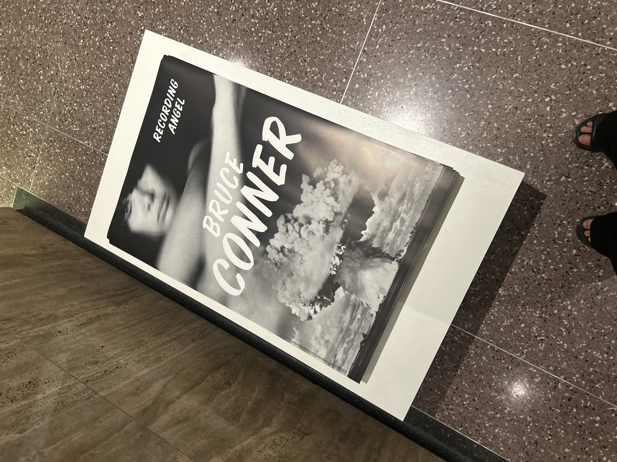 A close-up shot captures a book, entitled "Bruce Conner: Recording Angel", placed on the floor. The book's cover is black and white, featuring a person's face and a mushroom cloud. The book rests on a white surface. The floor is patterned with small, multi-colored spots. Parts of a person's feet, wearing sandals, are visible in the upper right corner. The photograph was taken inside a building, located in the city of Los Angeles, United States of America.