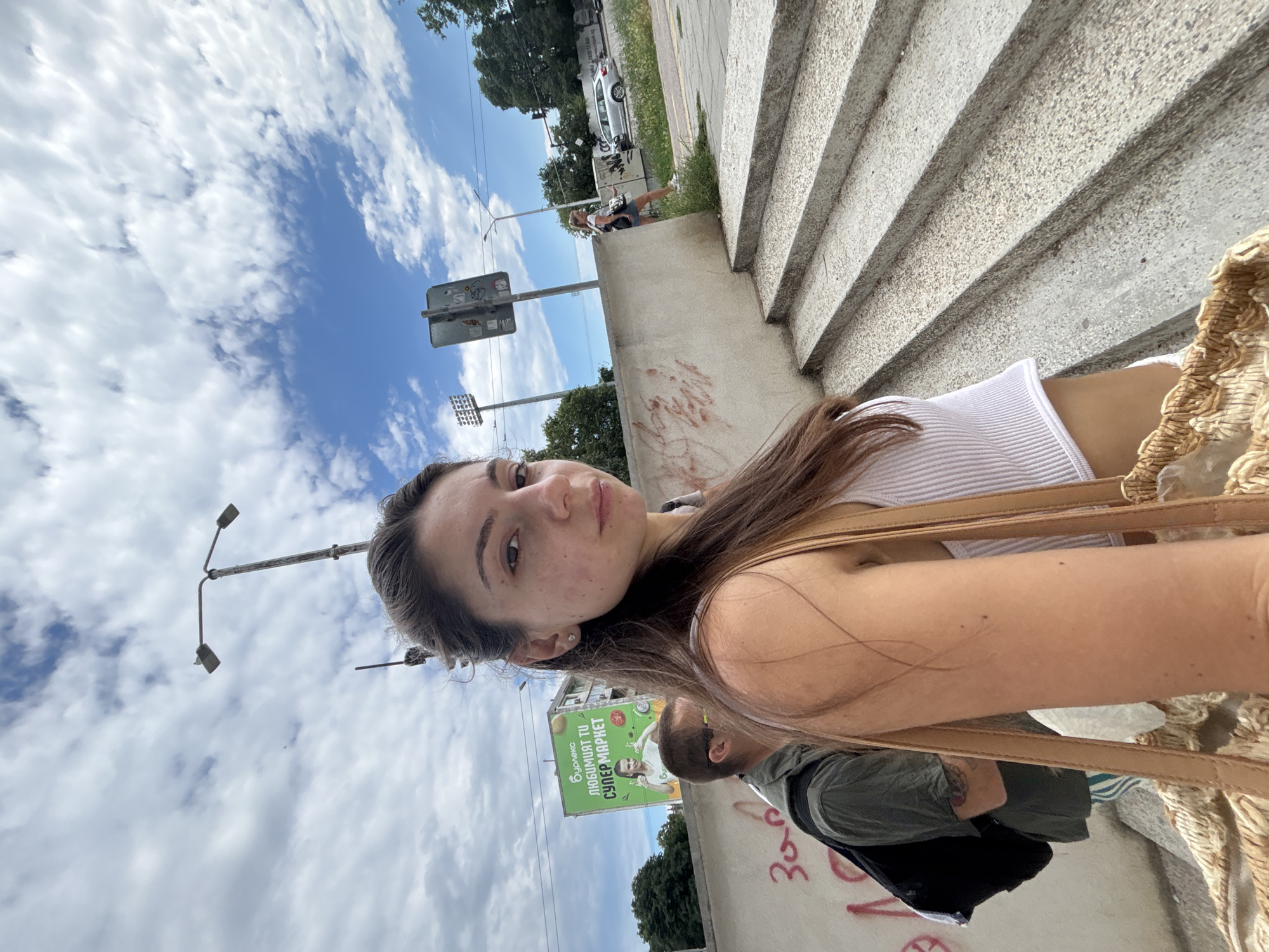 A young woman, facing forward, is captured in a selfie on a set of outdoor concrete stairs in Varna, Bulgaria. She has long brown hair, pulled back, and is wearing a white ribbed tank top and white shorts. She carries a large, woven straw bag over her shoulder. To her left, another person with short brown hair and glasses, wearing a green shirt and a backpack, is partially visible. Behind them, a large green billboard displays text and an image of a woman giving a thumbs-up. The background shows a cloudy sky with blue patches, trees, a street light, traffic signals, and a silver car on a road. The concrete wall next to the stairs is marked with graffiti.