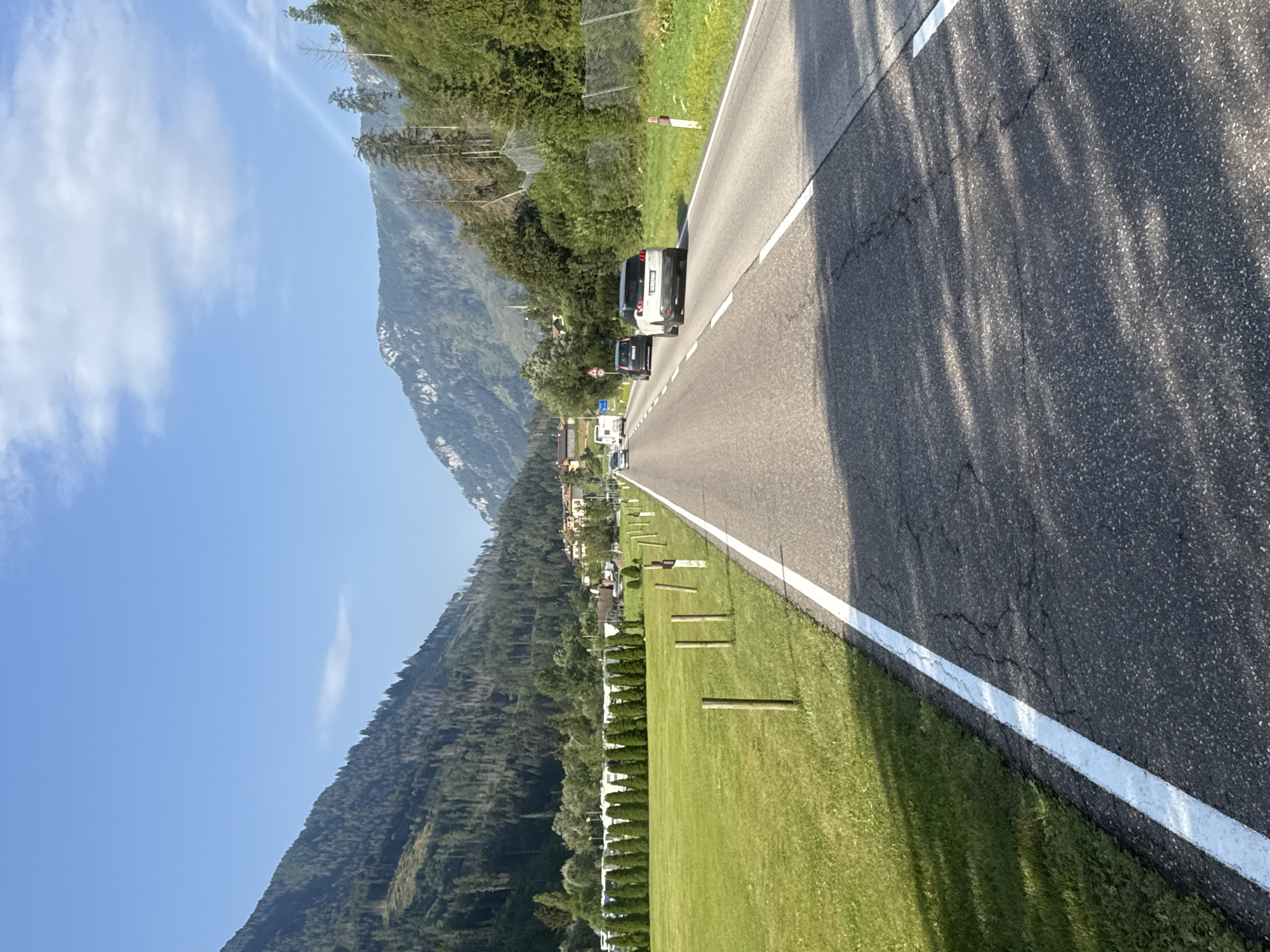 The image depicts a scenic view of a road in Moena, Italy, a town nestled in the Trentino region. The road, paved with asphalt and marked with white lines, stretches into the distance, flanked by lush green fields on the left and a dense forest on the right. The landscape is dominated by majestic mountains that rise in the background, their slopes covered with trees. The sky above is a clear blue, with a few wispy clouds adding texture to the scene. Several cars are visible on the road, suggesting ongoing traffic. The overall atmosphere is one of tranquility and natural beauty, with the human presence subtly integrated into the picturesque environment.