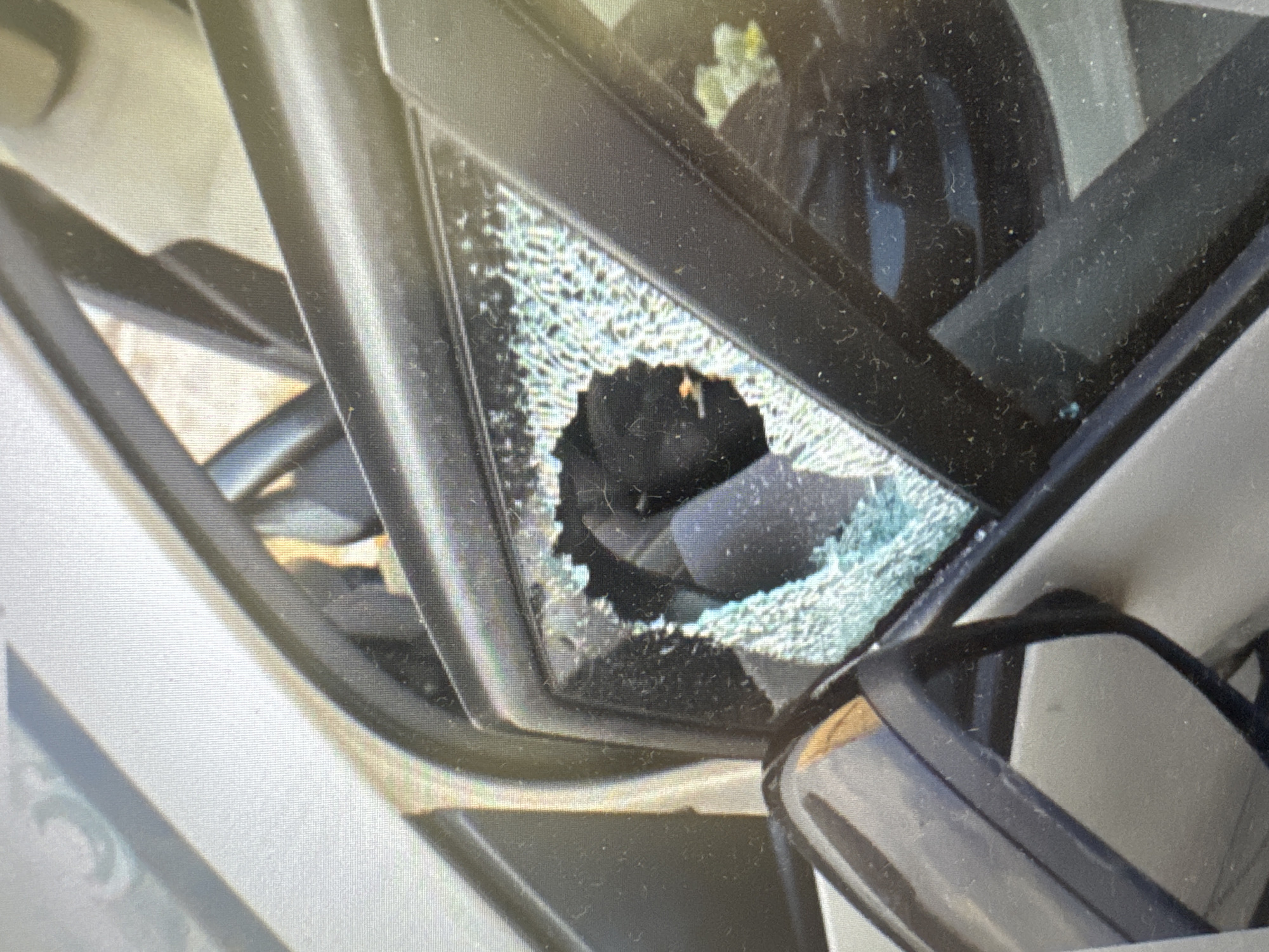 The image shows a close-up view of a car's side mirror and a broken window. The window is shattered, with glass fragments visible around a large hole in the center. The car appears to be a light color, possibly silver or white. The interior of the car is partially visible, showing dark-colored seats and trim. The scene is outdoors, with some foliage visible through the broken window. The time of day is not discernible, nor is the weather. There are no people or visible text in the image. The setting is likely a residential or public area where a car would be parked. The damage to the window suggests a potential break-in or vandalism. The provided location context is Murcia, Spain, but there are no specific visual cues in the image to confirm this.