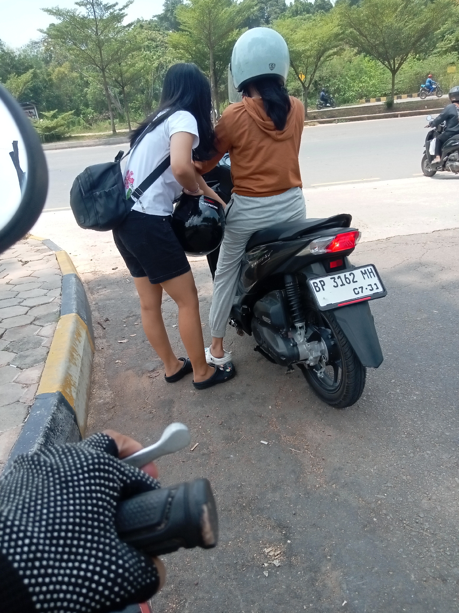 The image depicts two young women on a motorcycle parked on the side of a road in Batam, Indonesia. The woman in front wears a light blue helmet and an orange hoodie with grey sweatpants. The woman behind her, facing away from the camera, has long black hair and wears a white t-shirt, black shorts, and black Crocs. She is holding a black helmet with "HONDA" written on it. A black backpack is slung over her shoulder. The motorcycle is black with a license plate that reads "BP 3162 MH 07-31". The environment is an urban or suburban setting with a paved road, a sidewalk with hexagonal tiles, and trees and foliage in the background. The perspective suggests the photo is taken from another motorcycle, with a gloved hand holding the handlebar in the foreground.