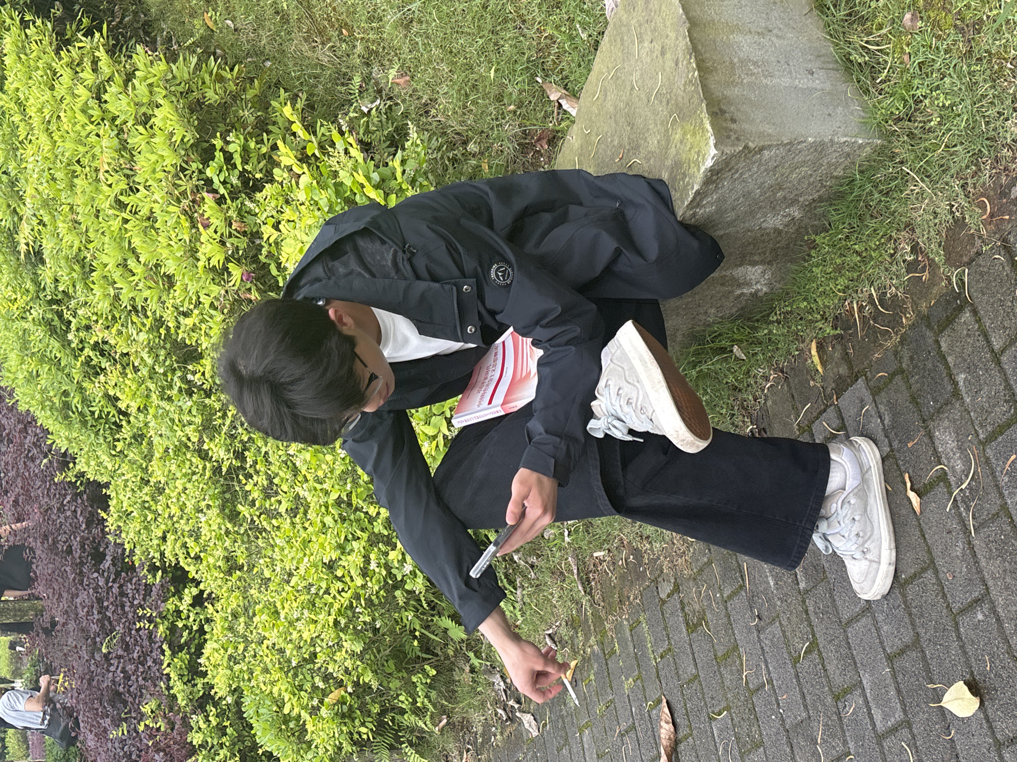 A person is crouching outdoors in a park-like setting. The individual has dark hair and is wearing glasses, a white t-shirt, a black jacket, and black pants. They are holding a book with red and white text and illustrations in one hand and a smartphone in the other. They are also holding a cigarette butt in their hand. They are wearing white sneakers, one foot resting on a concrete block. The person is positioned on a brick pathway with patches of grass and leaves. Lush green bushes and darker purple foliage are visible in the background. The location is suggested to be Jiangjin, China.