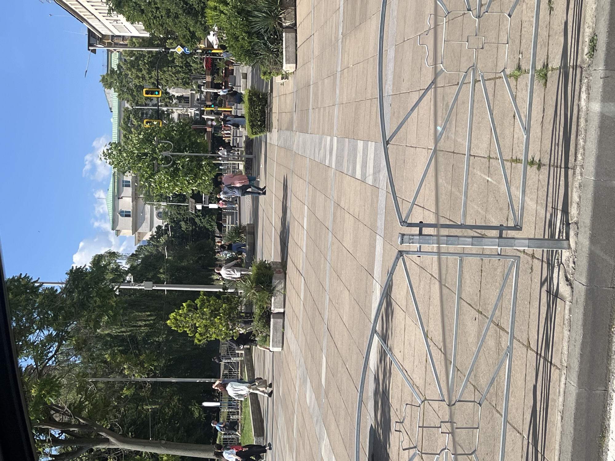 A street scene in an urban environment, likely on a sunny day with a clear blue sky. In the foreground, a silver metal fence with an ornate decorative pattern is visible, casting shadows on the beige tiled pavement below. The pavement has a geometric pattern with alternating light and dark grey strips.
Further into the scene, several people are walking across the square, some individually and some in small groups. They are dressed in casual summer clothing. Trees with lush green foliage are scattered throughout the area, providing shade. In the mid-ground, there are large planters with green bushes and small trees. Beyond the trees and planters, buildings with light-colored facades and green roofs are visible, along with traffic lights and street signs. The architecture suggests a European city.