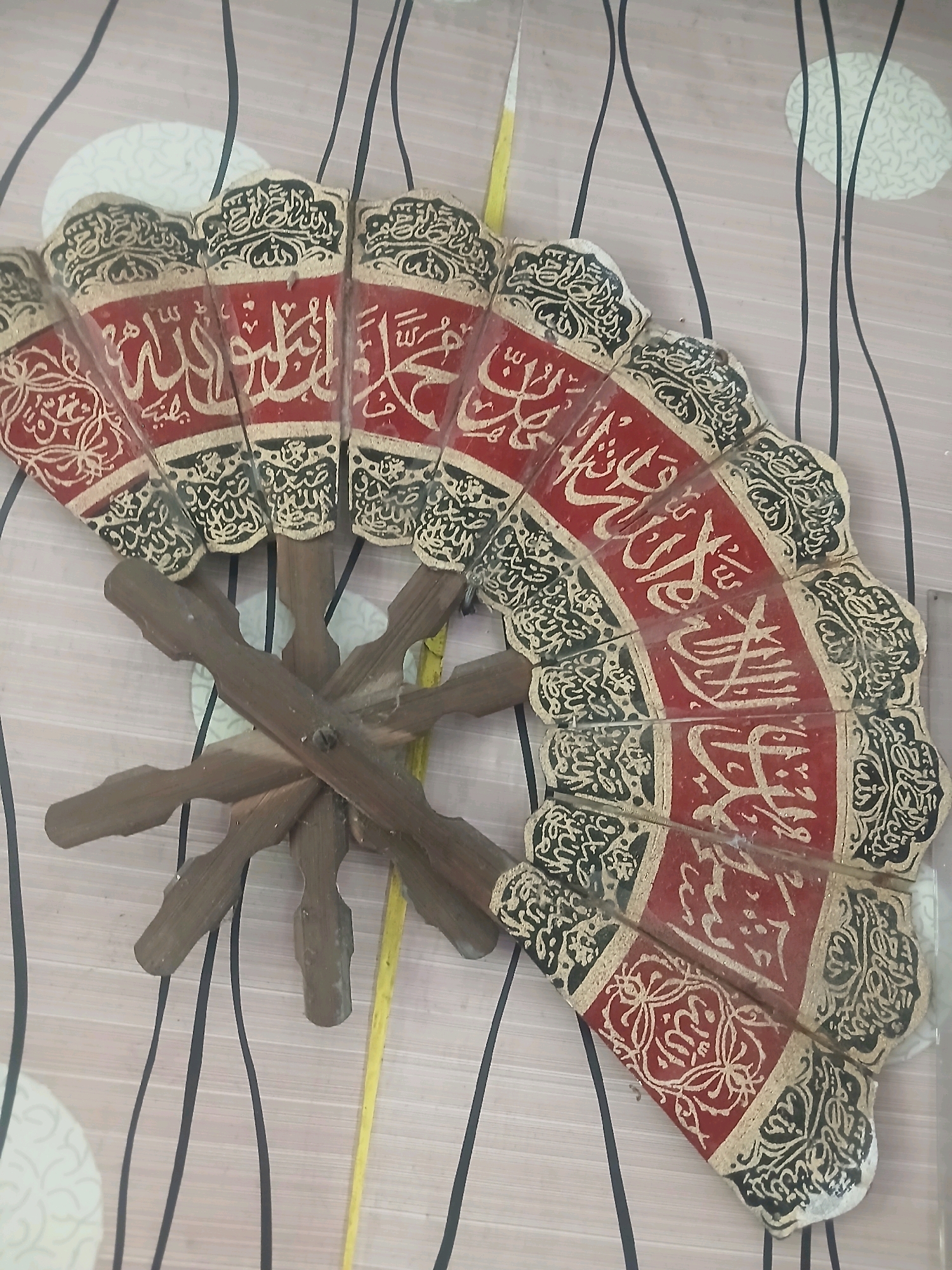 A wooden fan decorated with Islamic calligraphy is displayed against a backdrop of a striped wallpaper. The fan is made of several panels, each featuring Arabic script in gold on a red background, bordered by decorative black and gold patterns. The fan is held open by a wooden cross-shaped stand. The wallpaper has a pinkish-beige base with thin, dark grey, wavy vertical lines and a circular pattern of lighter beige with a textured design. A yellow measuring tape is visible partially in the frame, alongside the fan. The overall scene suggests a decorative item placed on a wall.