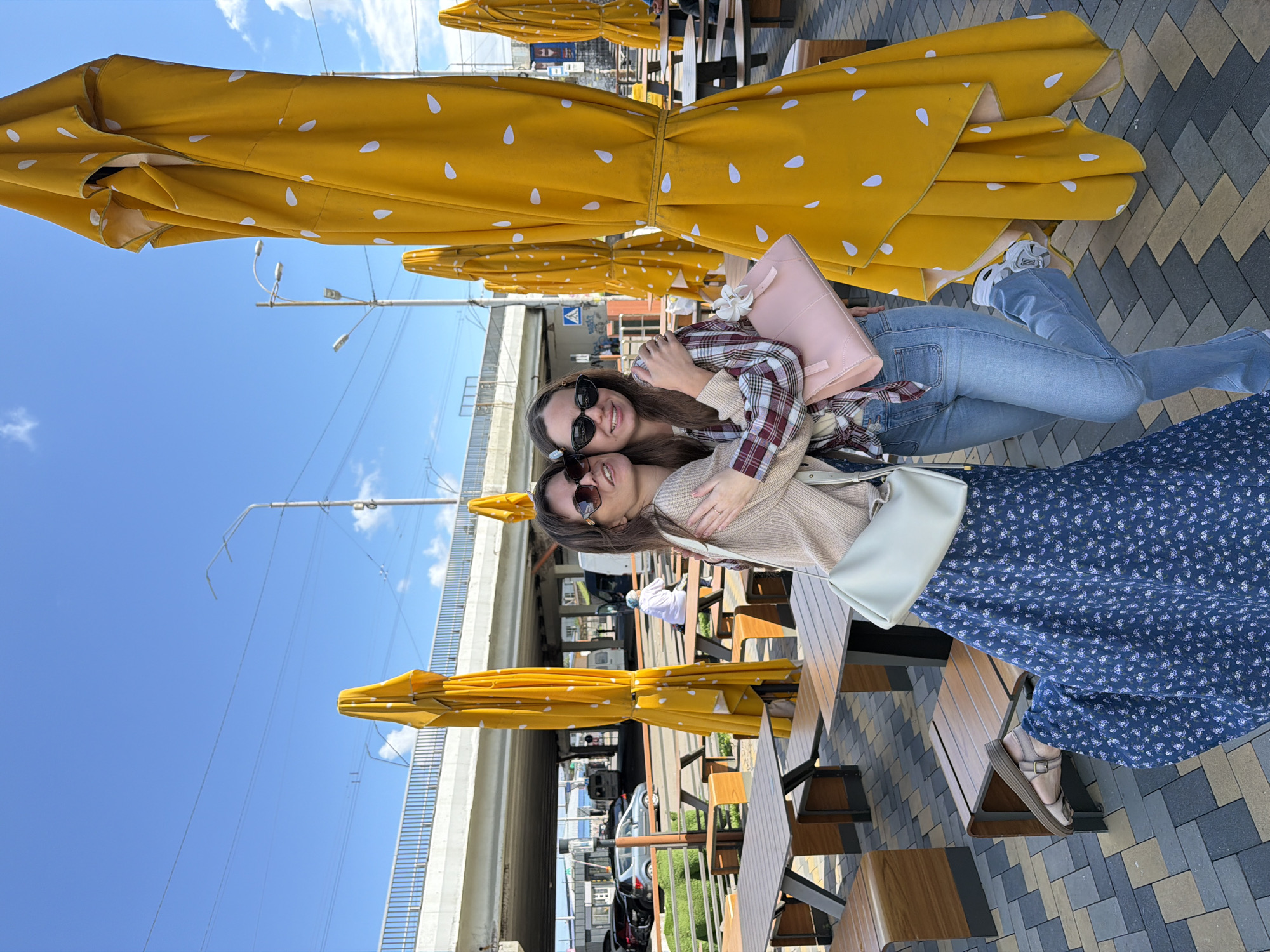 Two young women are embracing outdoors in an urban setting with a clear blue sky. The woman on the left wears a long, dark blue floral dress, a light beige cardigan, and sunglasses. She has a light beige crossbody bag. The woman on the right is wearing a plaid shirt, light blue jeans, and white sneakers, and is holding a pink handbag. They are standing in front of several large, closed yellow umbrellas with a white speckled pattern. In the background, there are outdoor tables and benches, a pedestrian walkway, a road with cars, and an overpass with power lines. The pavement is made of gray and brown interlocking blocks. The presence of the overpass, a common feature in Kyiv, suggests the location is likely within the city.
