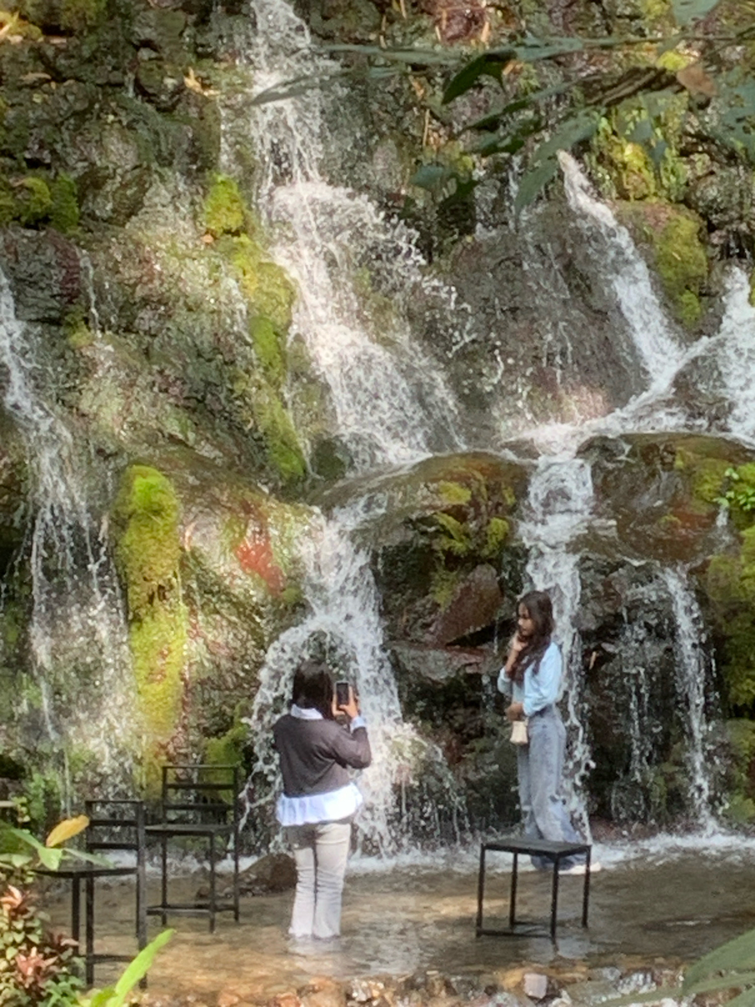 The image captures a scene at the Pronojiwo waterfall in Pronojiwo, Indonesia. Two young women are present. One woman, on the right, is posing with her hand near her chin, looking away from the camera, and is wearing a light blue long-sleeved shirt and grey pants. The other woman, on the left, is facing away from the camera and taking a picture with her phone. She is wearing a dark sweater over a white blouse and grey pants. Both women are standing in shallow water at the base of the waterfall. The waterfall cascades down a rocky, moss-covered cliff face. In the foreground, to the left, are two dark metal chairs and a small table. The environment appears lush and natural, with greenery surrounding the waterfall.