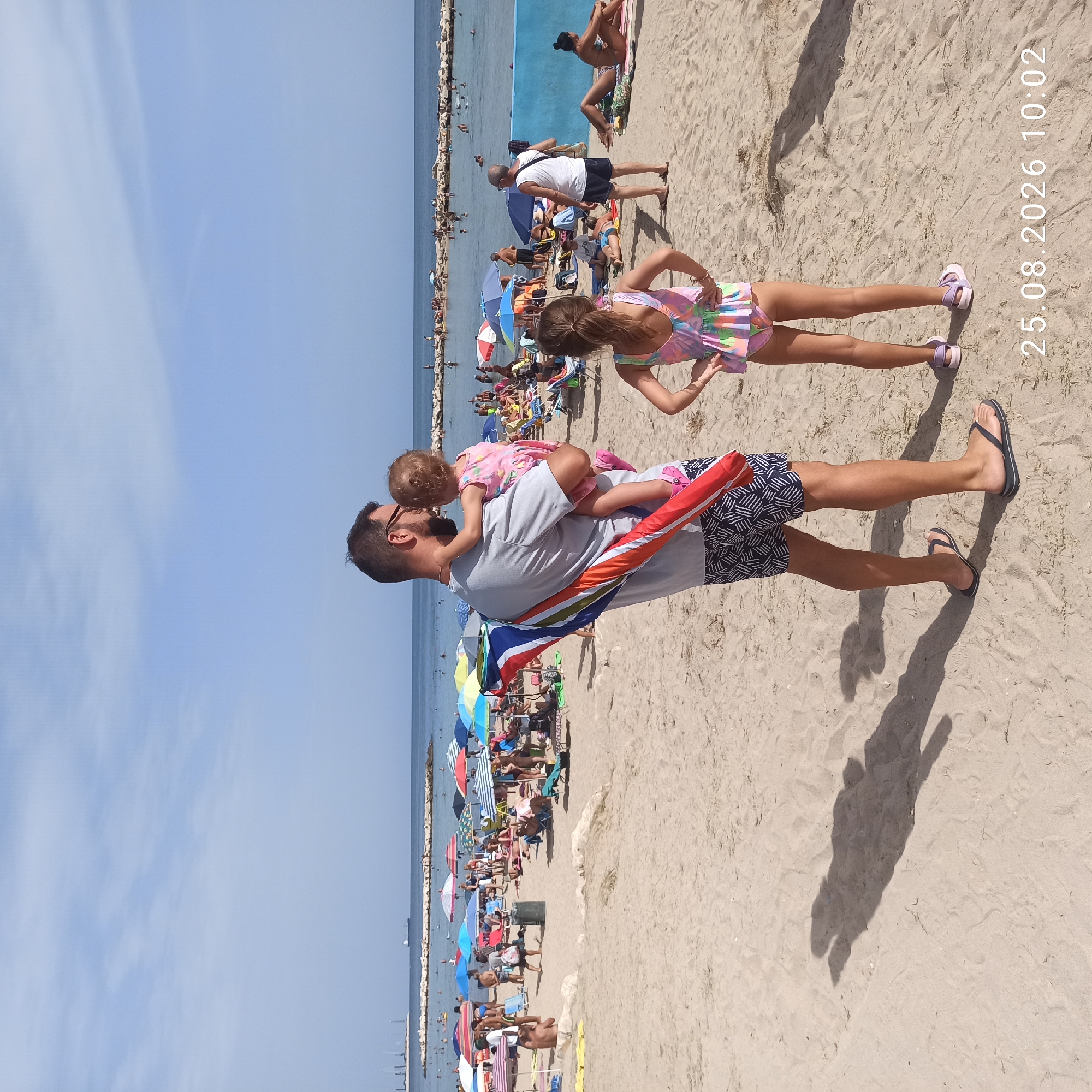The image shows a beach scene in Bari, Italy, on August 25, 2026, at 10:02 AM. The sand is a light beige, and the water is a calm blue. The sky is clear with a few wispy clouds.
In the foreground, a man is holding a toddler. He is wearing sunglasses, a grey t-shirt, and patterned shorts. The toddler is wearing a pink dress. To the right of the man, a young girl is standing with her hands on her hips. She is wearing a colorful swimsuit.
In the background, many people are on the beach, some sunbathing and others swimming in the sea. Numerous beach umbrellas are set up, providing shade. A ship is visible in the distance on the horizon, and a breakwater is present along the coastline. The overall impression is a busy, sunny day at the beach.