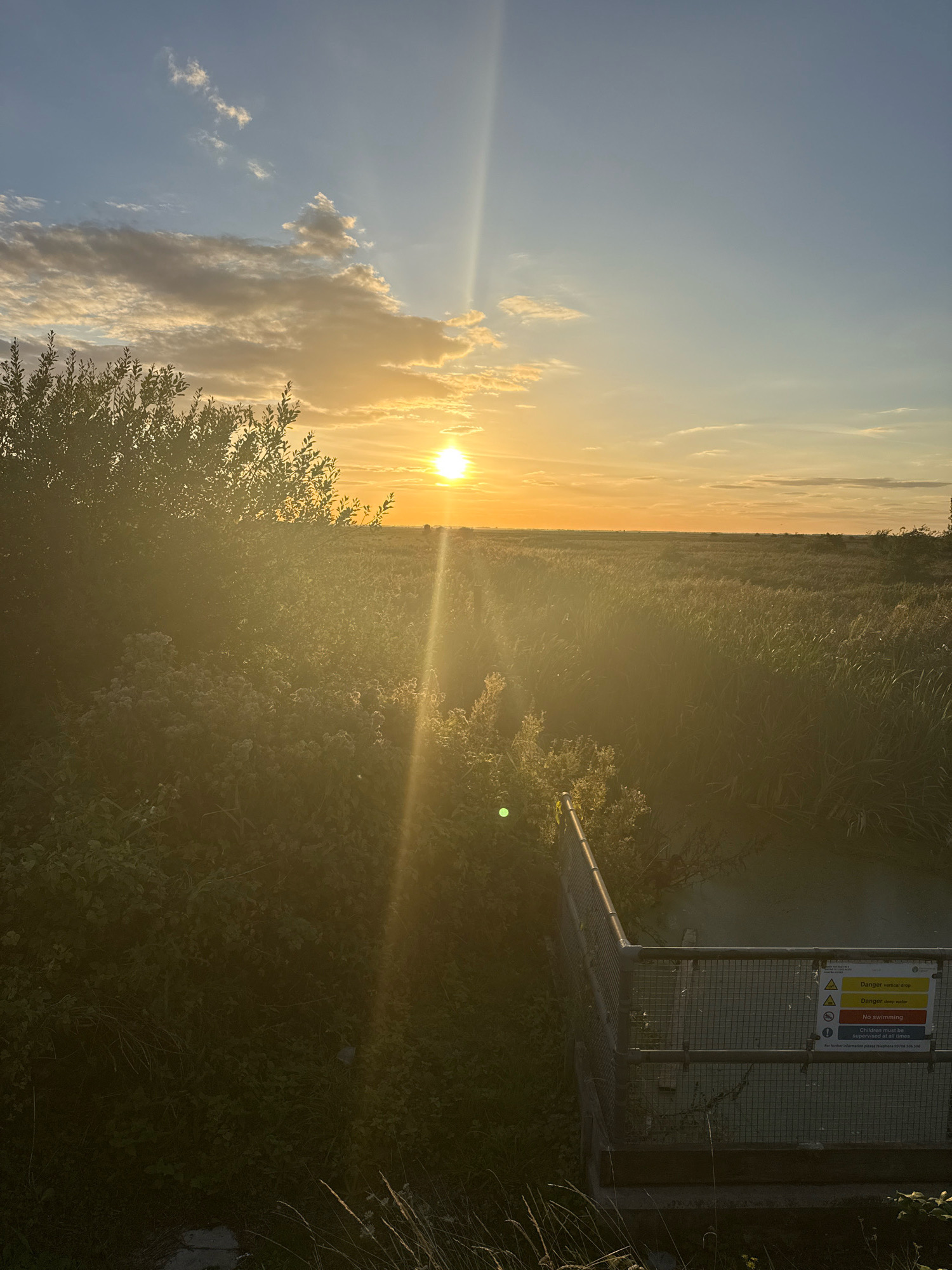 The image depicts a sunset over a body of water, likely a canal or drainage ditch, in Great Yarmouth, United Kingdom. The sun is low on the horizon, casting a warm glow across the sky and reflecting off the water. Tall reeds and grasses surround the water, with a metal fence and a warning sign indicating "Danger: vertical drop," "Danger: deep water," and "No swimming." The sign also states that "Children must be supervised at all times." The overall scene suggests a natural area within the urban environment of Great Yarmouth.
