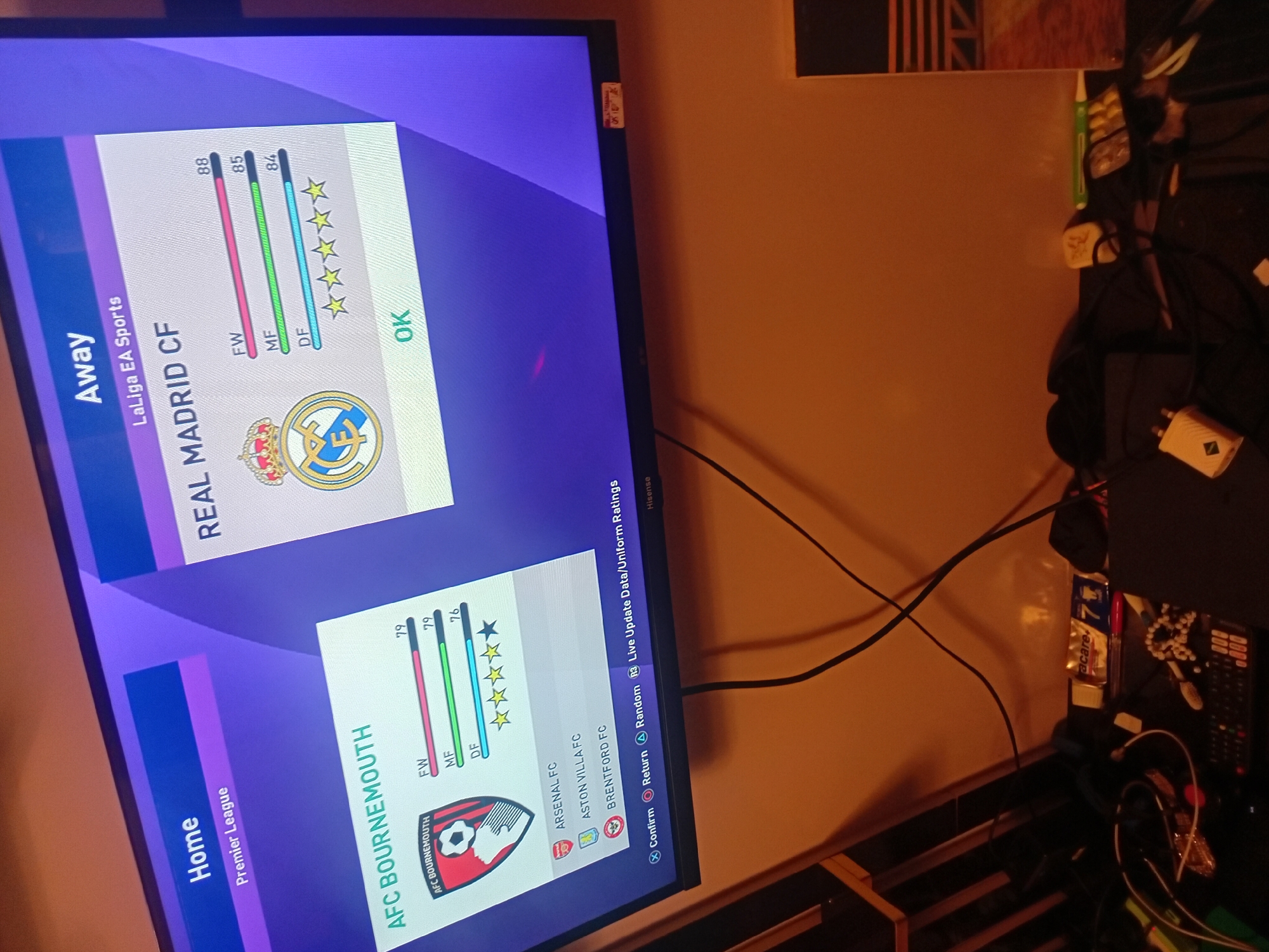 A television screen dominates the left side of the frame, angled as if viewed from above and to the left. It displays a video game interface, likely a football (soccer) simulation, showing team statistics and selection options. The game is comparing "REAL MADRID CF" as the "Away" team from "LaLiga EA Sports," with forward (FW) 88, midfield (MF) 85, and defense (DF) 84 ratings, earning five stars. Below, "AFC BOURNEMOUTH" is the "Home" team from the "Premier League," with FW 79, MF 79, and DF 76 ratings, earning three and a half stars. Other team names visible include "ARSENAL FC," "ASTON VILLA FC," and "BRENTFORD FC." Control prompts like "Confirm," "Return," "Random," and "R3 Live Update Data/Uniform Ratings" are also visible. The TV's brand, "Hisense," is written on the bottom right bezel.
The room appears dimly lit with a warm, orange glow on the wall behind the TV, suggesting artificial lighting or a warm-toned environment, possibly evening or night. A tangle of black cables runs across the wall and onto a cluttered surface to the right of the television. This surface holds various items including a remote control, a white power adapter, a green pen, blister packs of pills, and a clear plastic container labeled "acare+." A small sticker with obscured text is affixed to the top right edge of the TV bezel. The overall scene depicts a casual indoor setting, likely a living room or bedroom, focused on gaming.