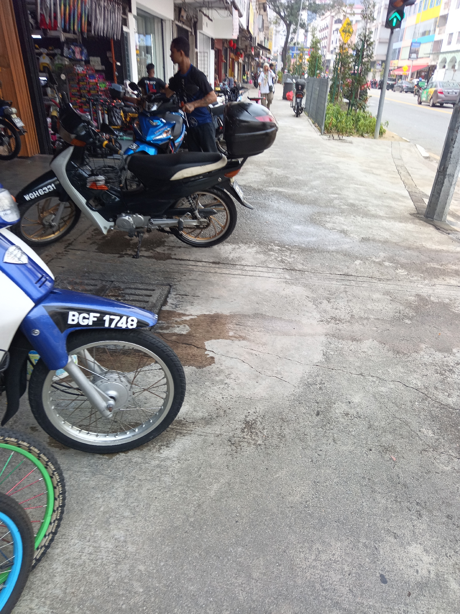 This image depicts a street scene in Kuala Lumpur, Malaysia, specifically near a row of shops. In the foreground, a blue and white motorcycle with the license plate "BGF 1748" is parked on a concrete sidewalk. To its left, another motorcycle's wheel with a green and red spoke rim is visible, suggesting multiple motorcycles are present or parked nearby.
Further back, a man in a black shirt is interacting with a blue motorcycle. Behind him, a rack of colorful items, possibly motorcycle accessories, is displayed outside a shop. Another motorcycle, predominantly white and black, with the license plate "WGH 8331," is also visible.
The sidewalk shows signs of wear and tear, with some damp patches, indicative of recent rain or cleaning. Beyond the sidewalk, a street is visible with traffic, including cars and a motorcycle, and a traffic light displaying a green arrow. The background features multi-story buildings, some with colorful facades, and trees lining the street, characteristic of an urban environment in Kuala Lumpur. The overall atmosphere suggests a busy commercial area.