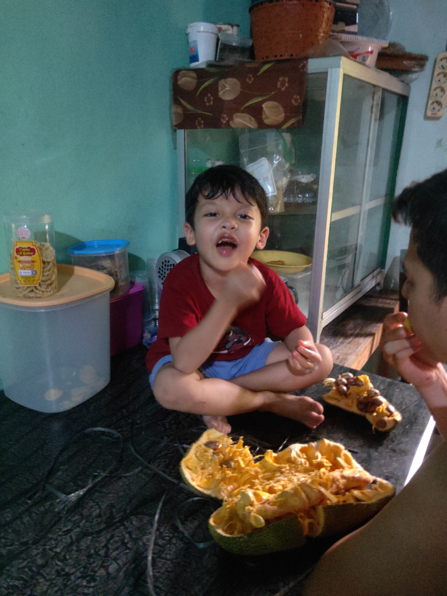 In a dimly lit room with teal walls, a young boy in a red shirt and blue shorts sits cross-legged on a black, textured surface. He is looking towards the camera with his mouth open, as if speaking or laughing, and his hands are held in front of him. To his right, a cut-open fruit, appearing to be a jackfruit or similar, lies on the surface, its bright yellow flesh and seeds exposed. Further to the right, part of an adult's arm and shoulder are visible, with their hand near their mouth, suggesting they are also interacting with the fruit. In the background, a glass-fronted cabinet stands against the wall, filled with various items, including containers and bags. A stack of plastic containers is also visible to the left of the boy, one of which holds a jar of snacks. The scene appears to be a domestic setting within the city of Sintang, Indonesia, possibly a kitchen or living area.