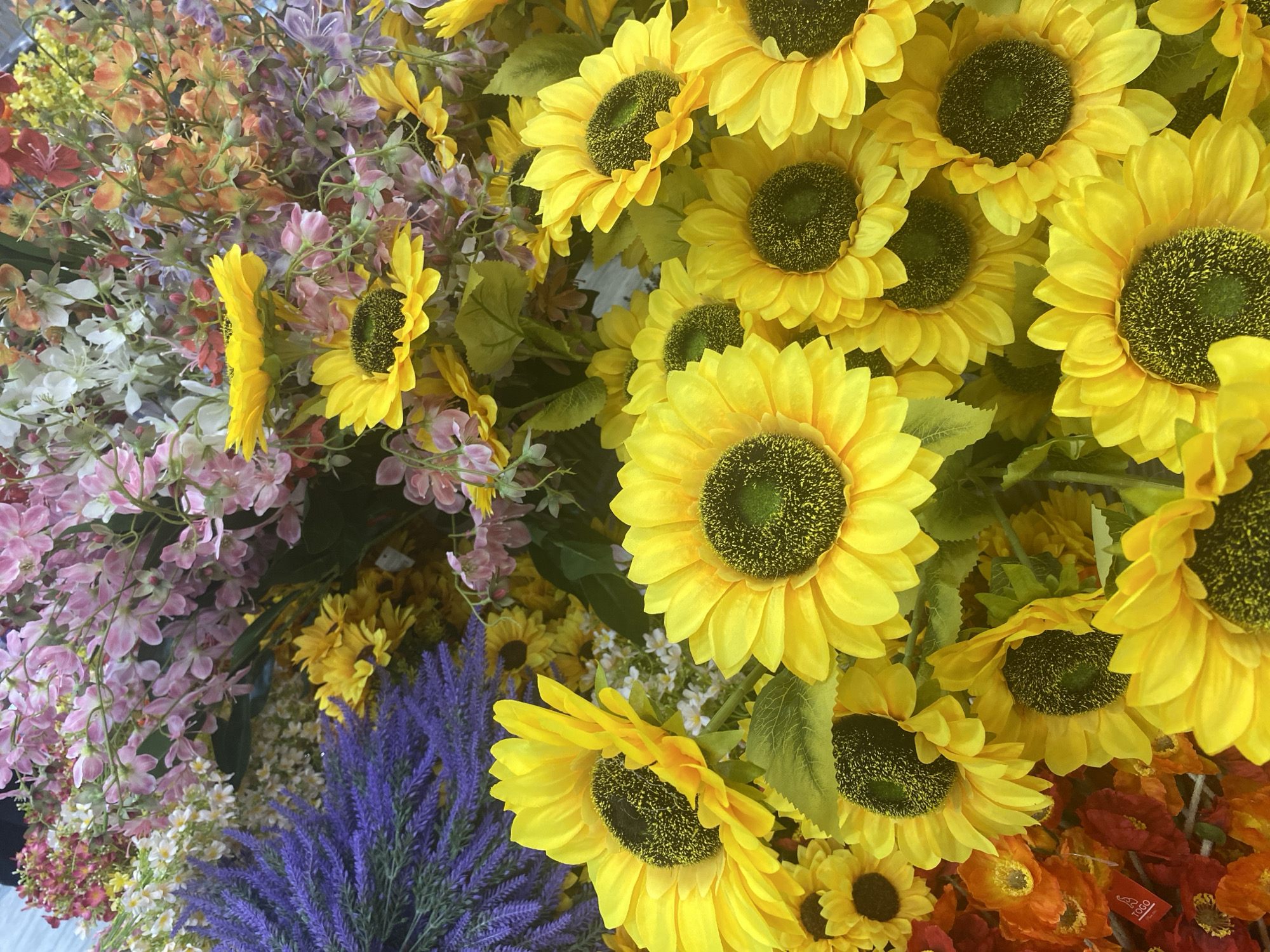 This is a close-up, overhead shot of a dense arrangement of artificial flowers. The foreground is dominated by numerous bright yellow sunflowers with dark brown centers. Behind the sunflowers, a variety of other artificial flowers are visible, including clusters of pink, light purple, and small white blossoms. In the upper left, a spray of lavender-like purple flowers adds another texture. The arrangement is tightly packed, suggesting it could be a display in a shop or part of a larger floral installation. There are no people or animals present. The setting appears to be indoors, possibly a florist shop or a market stall. The lighting is bright and even, highlighting the vibrant colors of the artificial flowers. No specific landmarks or urban features of Konya, Turkey, are discernible within this image.