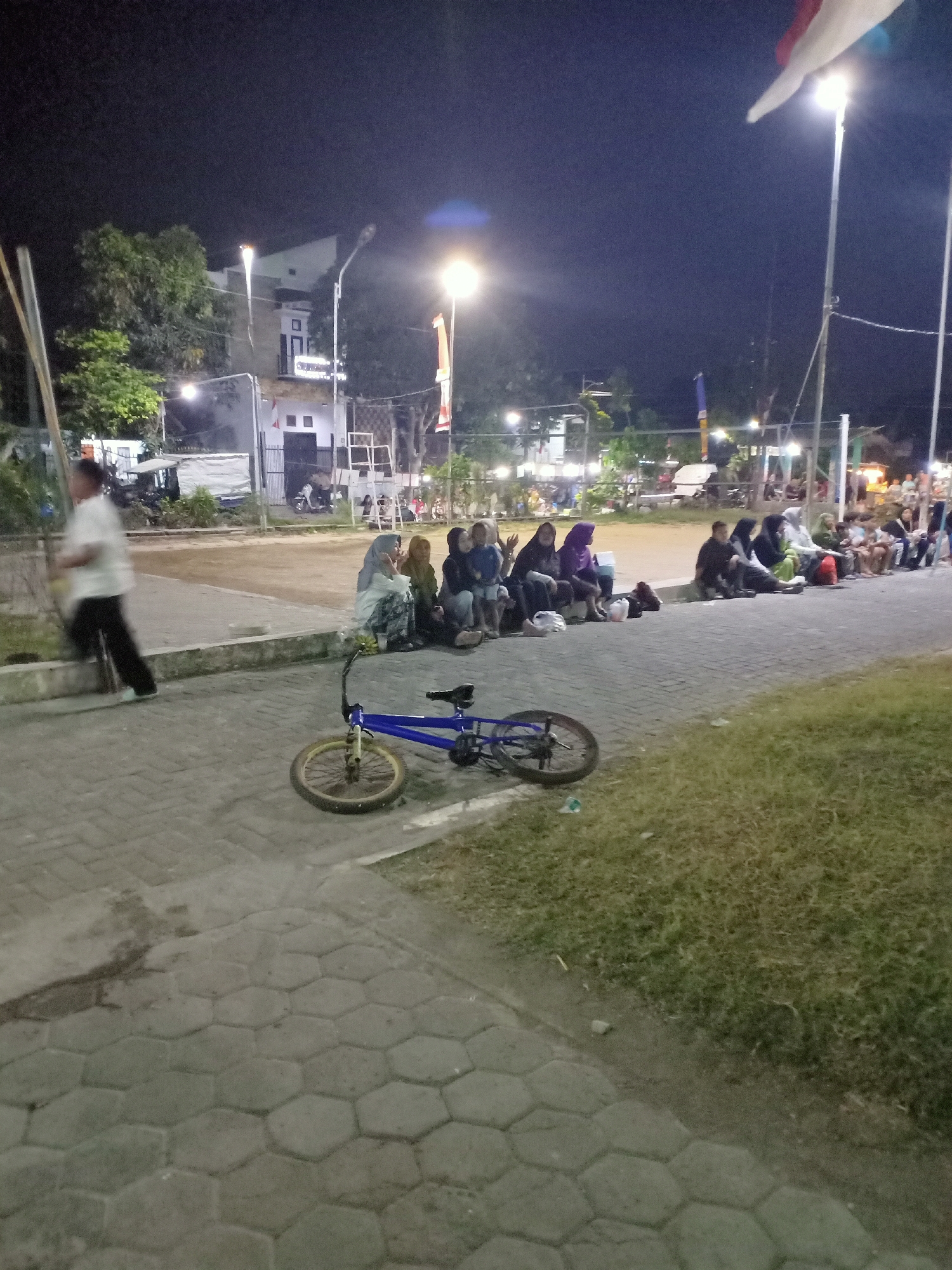 The image depicts an outdoor scene at night in Sumber Tlaseh, Indonesia. A group of people, some wearing hijabs, are seated on the ground along a paved walkway. In the foreground, a blue bicycle is lying on its side on the hexagonal patterned paving stones. A person in a white shirt and dark pants is walking away from the camera on the walkway. In the background, buildings with illuminated windows and streetlights are visible, along with trees and a sports court area. A flag is also visible on a pole. The overall lighting suggests it is evening or night, with artificial lights illuminating the area.