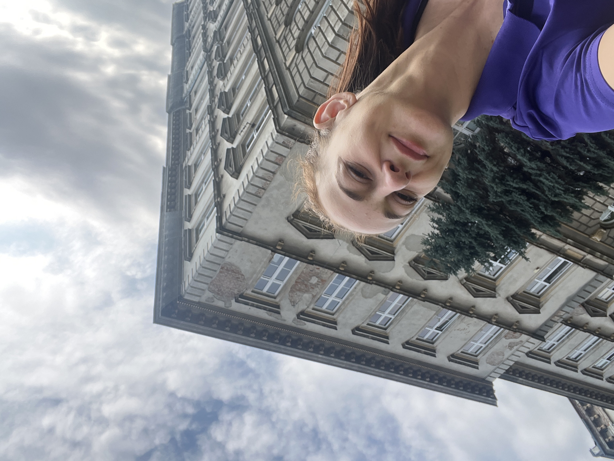 A woman is captured in a selfie, looking towards the camera with a slight smile. She has auburn hair and is wearing a vibrant purple collared shirt. Behind her, an aged, multi-story building with classical architectural details like decorative window surrounds and cornices dominates the frame. The building's facade appears worn, with some visible brickwork. A dense evergreen tree is situated in front of the building, partially obscuring some of its lower sections. The sky above is overcast with dramatic, cloud-filled patterns. The photograph is taken from a low angle, looking upwards towards the building and sky, with the woman positioned in the foreground.
