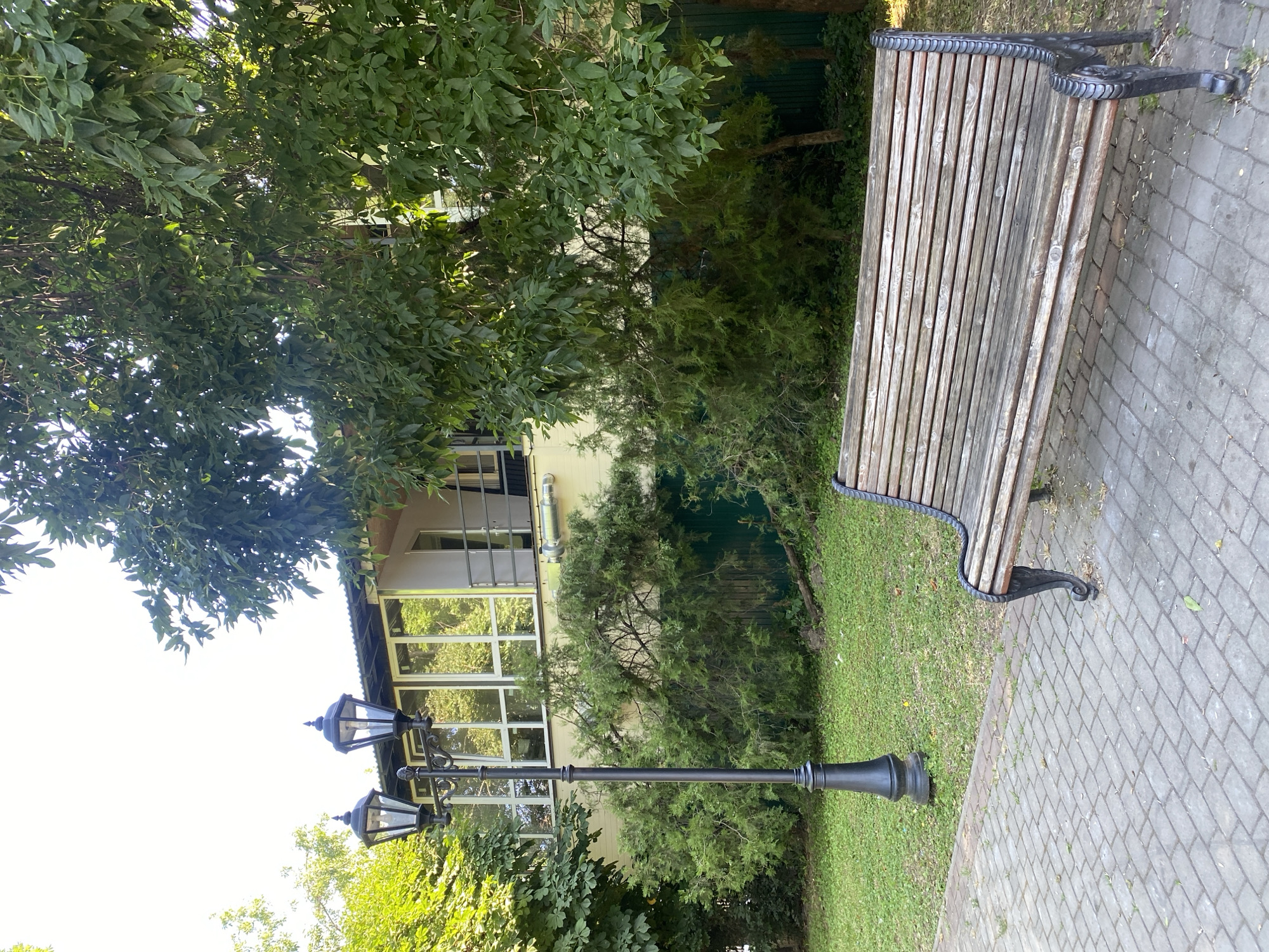 This is a vertical, eye-level outdoor shot of a park bench and a lamppost in front of a building. The scene is set in the Zapadny District of Russia. The bench is made of wood and black metal, and it is positioned on a paved walkway. To the left of the bench is a lamppost with two lamps, and behind it is a grassy area. The building in the background appears to be a two-story structure with large windows. There are trees and greenery surrounding the area, suggesting a peaceful, natural setting within the city. The sky is bright and clear, indicating a sunny day.