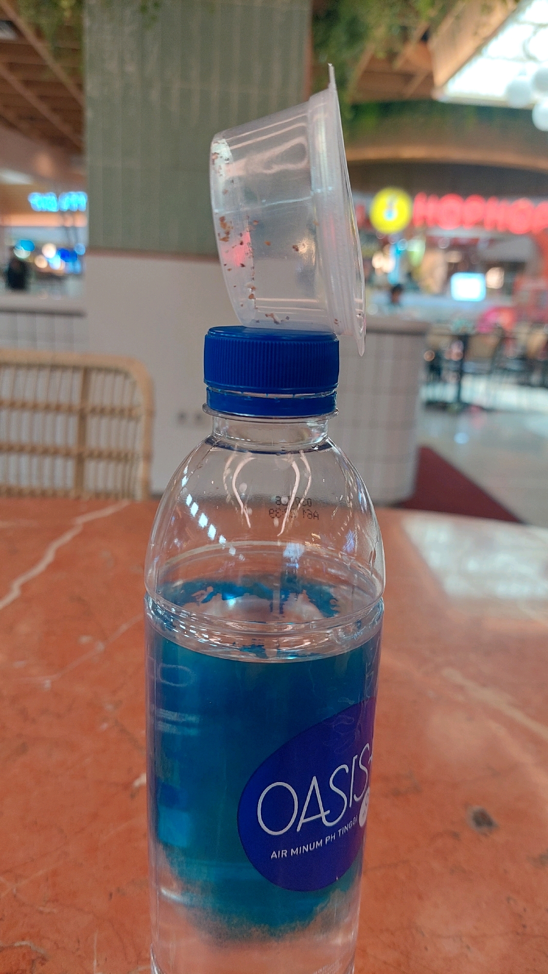 The image shows a close-up of a partially filled bottle of Oasis brand flavored water with a blue cap. A clear plastic lid, containing small brown particles, is balanced precariously on top of the bottle cap. The bottle has a blue label with the "OASIS" logo and text that reads "AIR MINUM PH TINGGI." The bottle is sitting on an orange-brown marble table. In the blurred background, there are elements of a public indoor space, possibly a food court or shopping mall. Signs with bright lighting are visible, including one that appears to have yellow lettering on a red background. Other elements in the background include light-colored furniture, green tiled walls, and hanging plants, suggesting a modern, commercial setting. Based on the Oasis branding and the common distribution of this product, the location is likely a public area in North Jakarta, Indonesia, such as a mall or restaurant within the city.