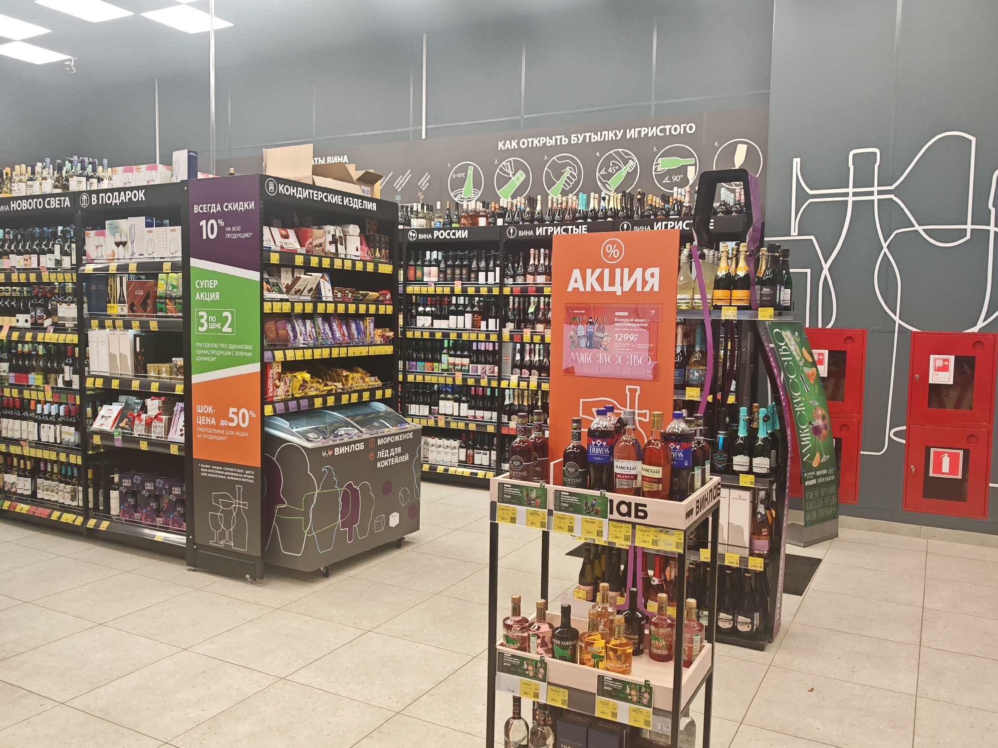 The image shows a retail store, likely a supermarket or liquor store, with shelves stocked with various alcoholic beverages, confectionery, and promotional displays. The overall environment suggests an indoor setting with artificial lighting.
On the left side of the image, there are tall shelves filled with wine bottles and boxes of confectionery. A prominent display board with the text "ВСЕГДА СКИДКИ 10% НА ВСЮ ПРОДУКЦИЮ" (Always discounts 10% on all products) is visible, along with a smaller banner promoting a "СУПЕР АКЦИЯ" (Super promotion) offering "3 по цене 2" (3 for the price of 2). To the right of these shelves, there is a refrigerated display case with the label "ВИП ЛОБ МОРОЖЕНОЕ ЛЕД ДЛЯ КОКТЕЙЛЕЙ" (VIP CLUB Ice cream Ice for cocktails).
The central and right parts of the image feature more shelves stocked with a variety of alcoholic beverages, including wine and spirits. A large orange promotional sign with the text "% АКЦИЯ" (Action) and images of bottles is centrally placed. Below this, a display stand showcases several bottles of spirits, with price tags visible. Further to the right, there are red fire safety cabinets mounted on the wall. The walls are painted in a neutral color and decorated with white line drawings of bottles and glasses.
The floor is tiled with a light-colored pattern. The ceiling has visible light fixtures. There are no people clearly visible in the frame. The general impression is a well-organized retail space with an emphasis on promotional offers for alcoholic products.
While the image itself does not contain explicit markers of its location in Moscow, Russia, the Cyrillic script on the signs and product labels strongly indicates a Russian-speaking region. Given the typical appearance of such retail environments, it is plausible that this is a store within a city like Moscow.