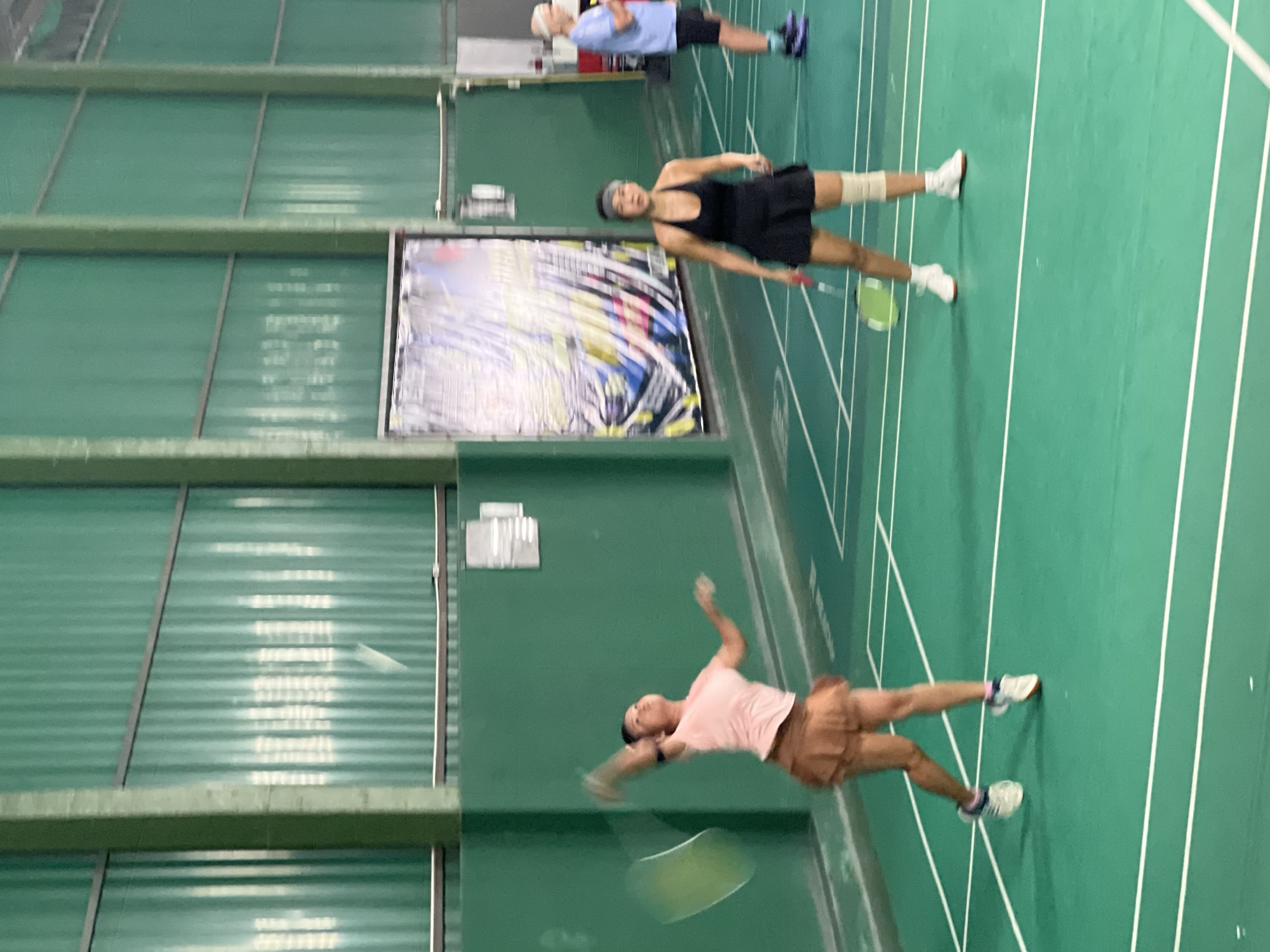 The image shows a badminton court, likely indoors due to the presence of an overhead ceiling and indoor lighting. There are three people in the frame. A woman in a pink shirt and brown shorts is captured in mid-swing, holding a badminton racket. Another woman, wearing a black dress and a bandage around one knee, stands on the opposite side of the court. Further in the background, another person, wearing a light blue shirt and dark shorts, stands near the back of the court. A large framed picture or banner is visible on the wall. The court has white lines marking the playing area on a green surface. The location is likely in บ้านบางรักษ์, Thailand.