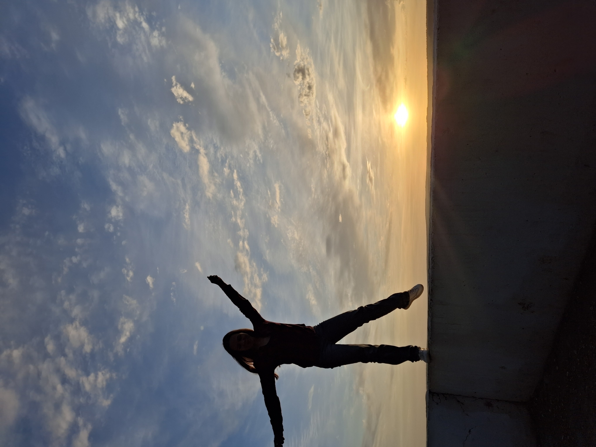 A photo, rotated 90 degrees counter-clockwise, depicts a person balanced on a concrete ledge against a backdrop of a partly cloudy sky and the setting sun. The person, silhouetted against the bright sky, is wearing pants and a long-sleeved top, with arms extended. The concrete ledge forms a sharp, dark contrast to the bright sky. The sky displays a gradient of blue and orange hues, indicating either sunrise or sunset. Based on the location information (Chaikovsky, Russia), this is likely a picture of the sunset over the concrete ledge.