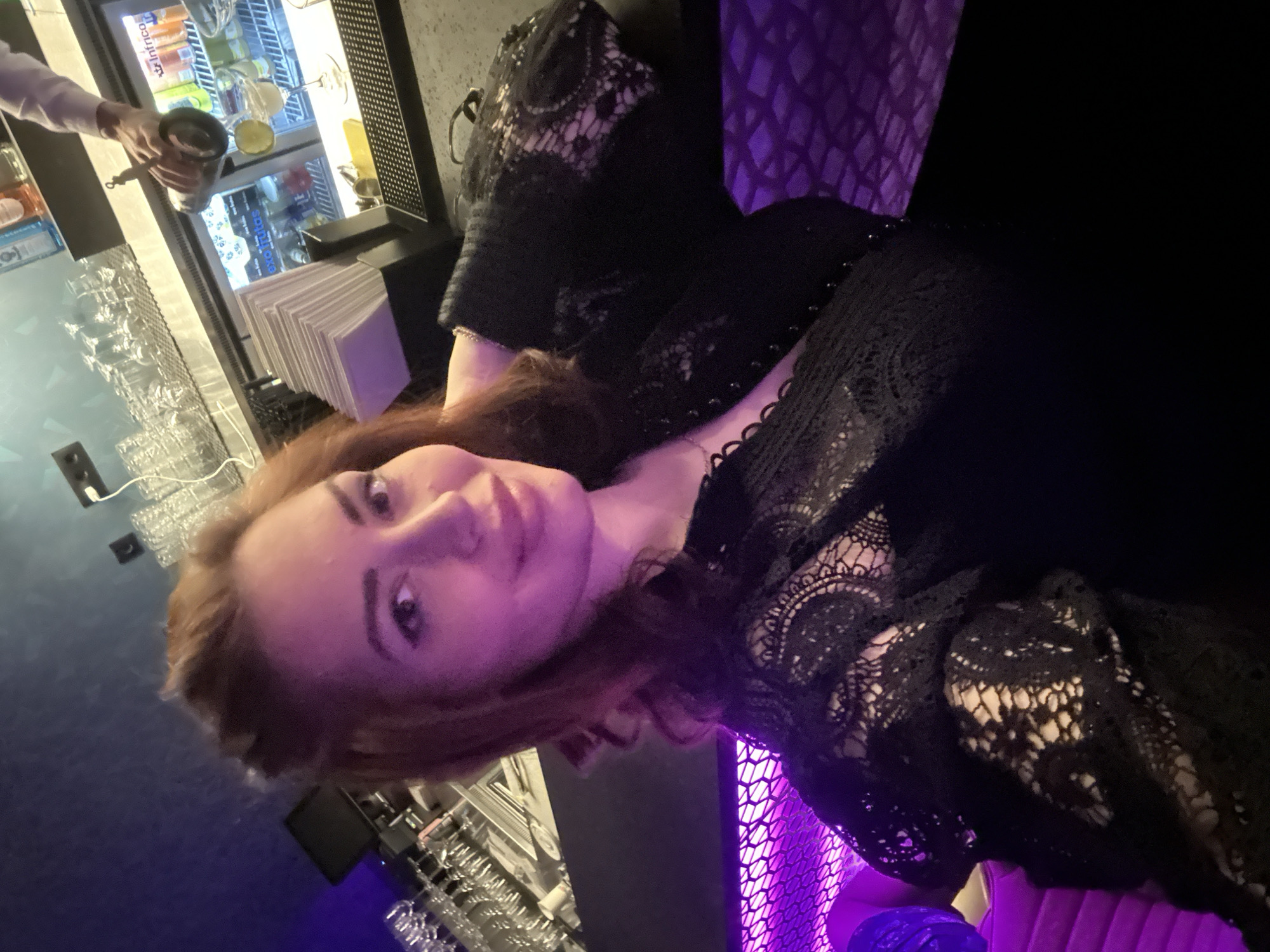 A woman with auburn hair and fair skin is the primary subject of a photograph. She is wearing a black, lace-embellished garment. The background includes a bar-like setting with stemware, napkins, a refrigeration unit containing beverage options, and lighting with blue and purple hues. An individual, partially visible, is holding a cocktail shaker near the bar. The location could be Kazanlak, Bulgaria.