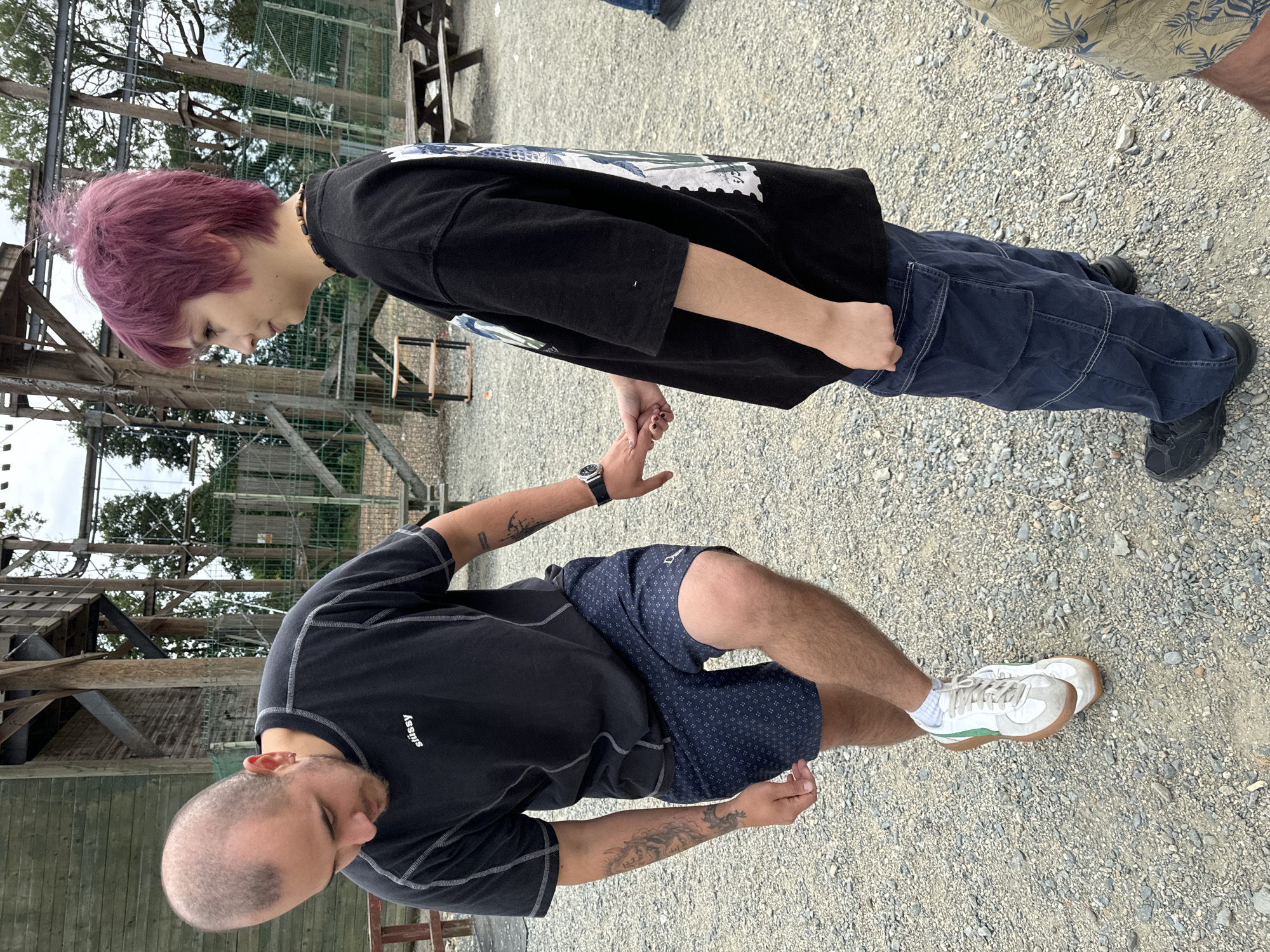 Two individuals are standing on a gravel surface outside what appears to be an adventure park or outdoor recreational facility. The person on the left, a male with a shaved head and tattoos on his arms, is wearing a black t-shirt with white stitching and blue patterned shorts. He is bending down and touching his right foot, which is clad in a white and green sneaker. The person on the right, a female with short purple hair, is wearing a black t-shirt with graphics on the back and dark blue cargo pants. Their hands are clasped together. In the background, there is a wooden structure resembling a climbing wall or ropes course, along with picnic tables and trees. The environment suggests an outdoor recreational area, likely within Courtown, Ireland.