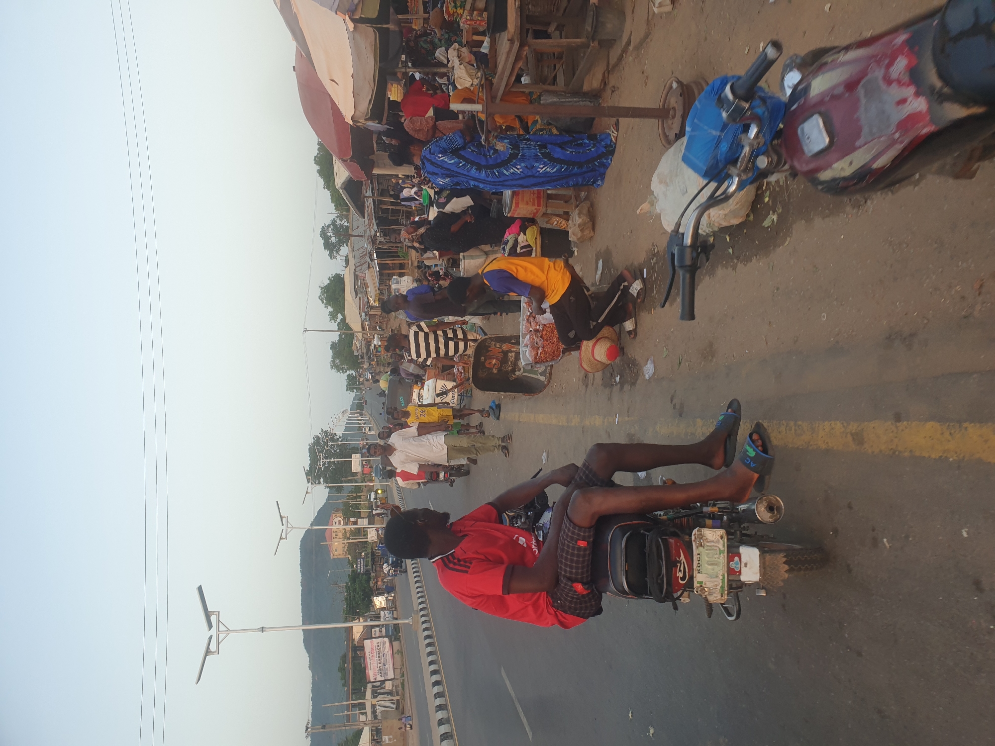 The image shows a street scene, seemingly a market area. In the foreground, a man is seated on a motorcycle on a road marked with a yellow line. He wears a red shirt, checkered shorts, and sandals. The motorcycle has a license plate labeled "KOGI" and appears to have some items strapped to it. In the background, there is a bustling market scene with several people and market stalls with awnings. The stalls are filled with various goods. Beyond the market and street, there are buildings and a distant mountain range. Overhead are street lights. The sky is overcast.