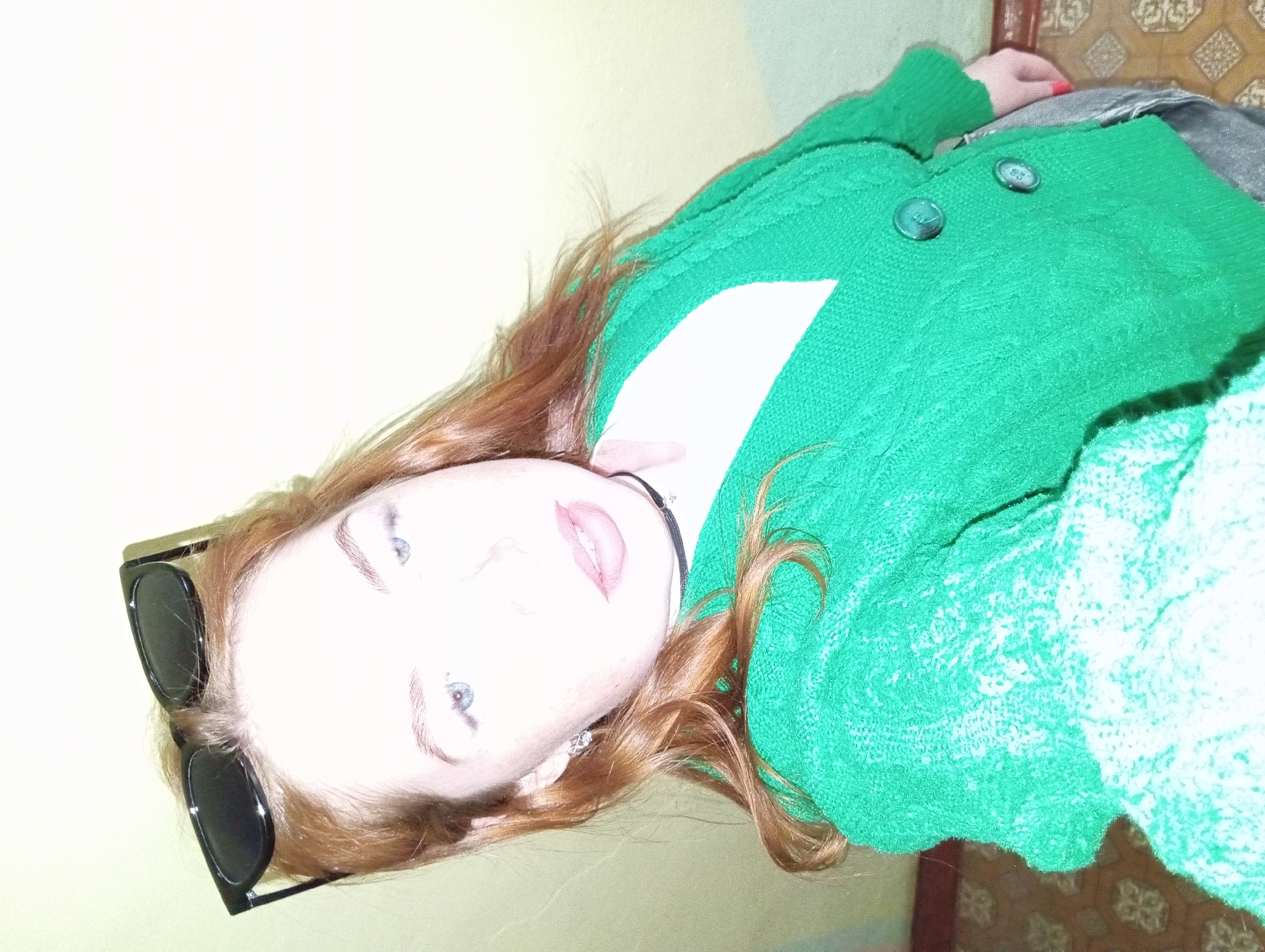 A person with fair skin and light blue eyes, wearing a black choker necklace and a green knit sweater with large buttons, is seen from a close, slightly upward angle. A pair of black sunglasses rests on their forehead. The background consists of a pale yellow wall on one side and a patterned surface or floor on the other. The location in the city of Yurgamysh, Russia, cannot be confirmed or inferred from the visual content of the image alone.