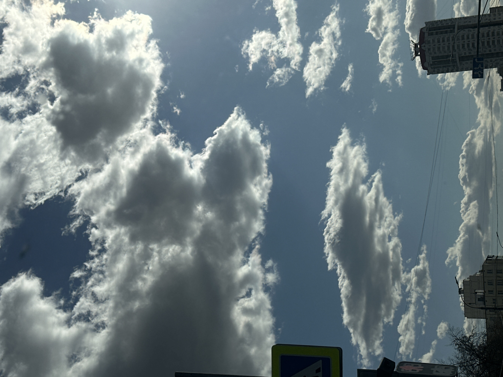 The image shows a view of the sky with clouds, taken from a ground-level perspective in Moscow, Russia. The sky is partially covered with cumulus clouds, with patches of blue sky visible between them. A tall building is visible on the right side of the frame, and a sign is visible at the bottom of the frame.