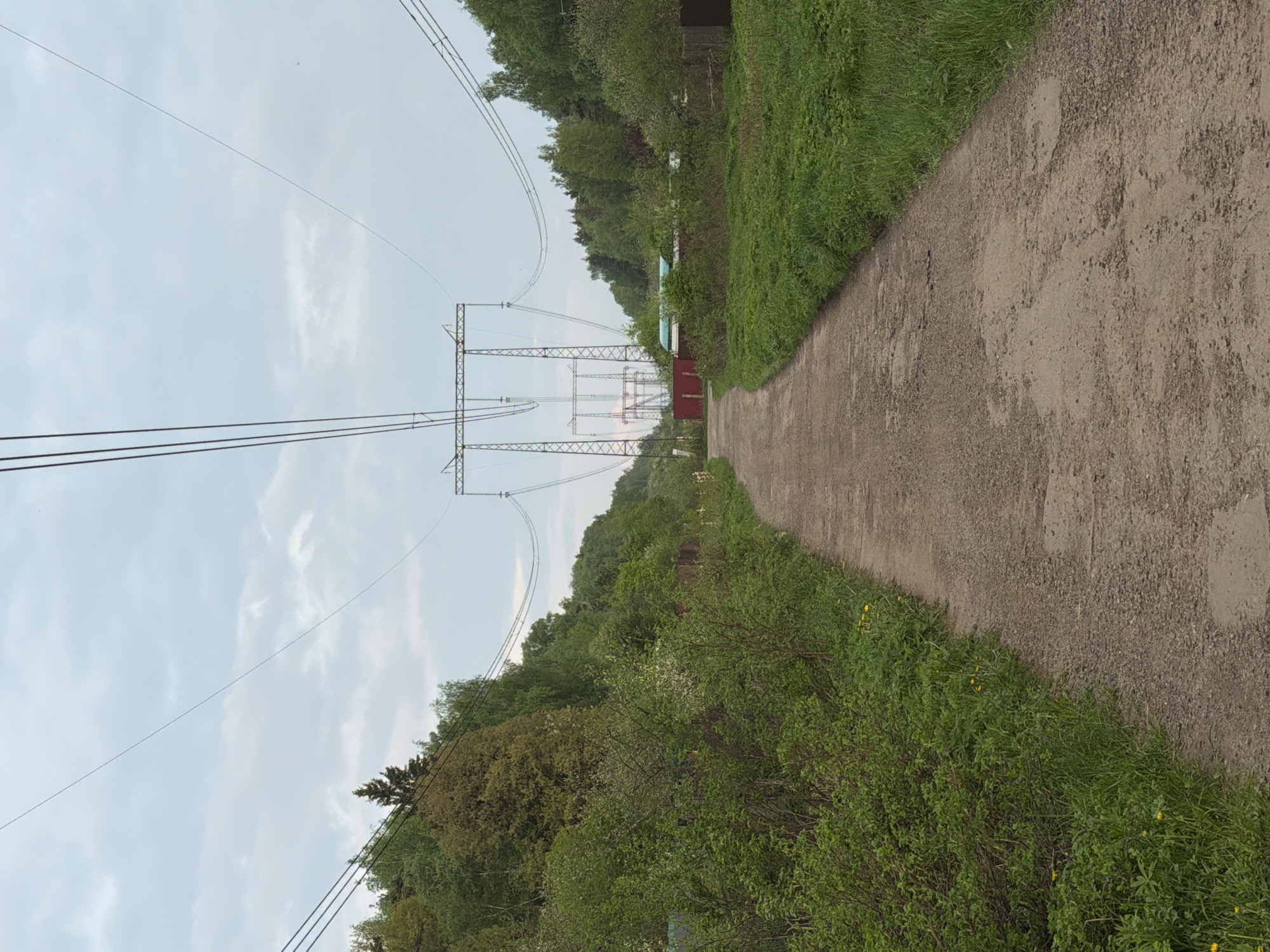 The image shows a dirt path running through a green area. Power lines and pylons are visible along the left side of the path. Vegetation lines both sides of the path, with a building visible in the distance. The location appears to be a rural or suburban area.