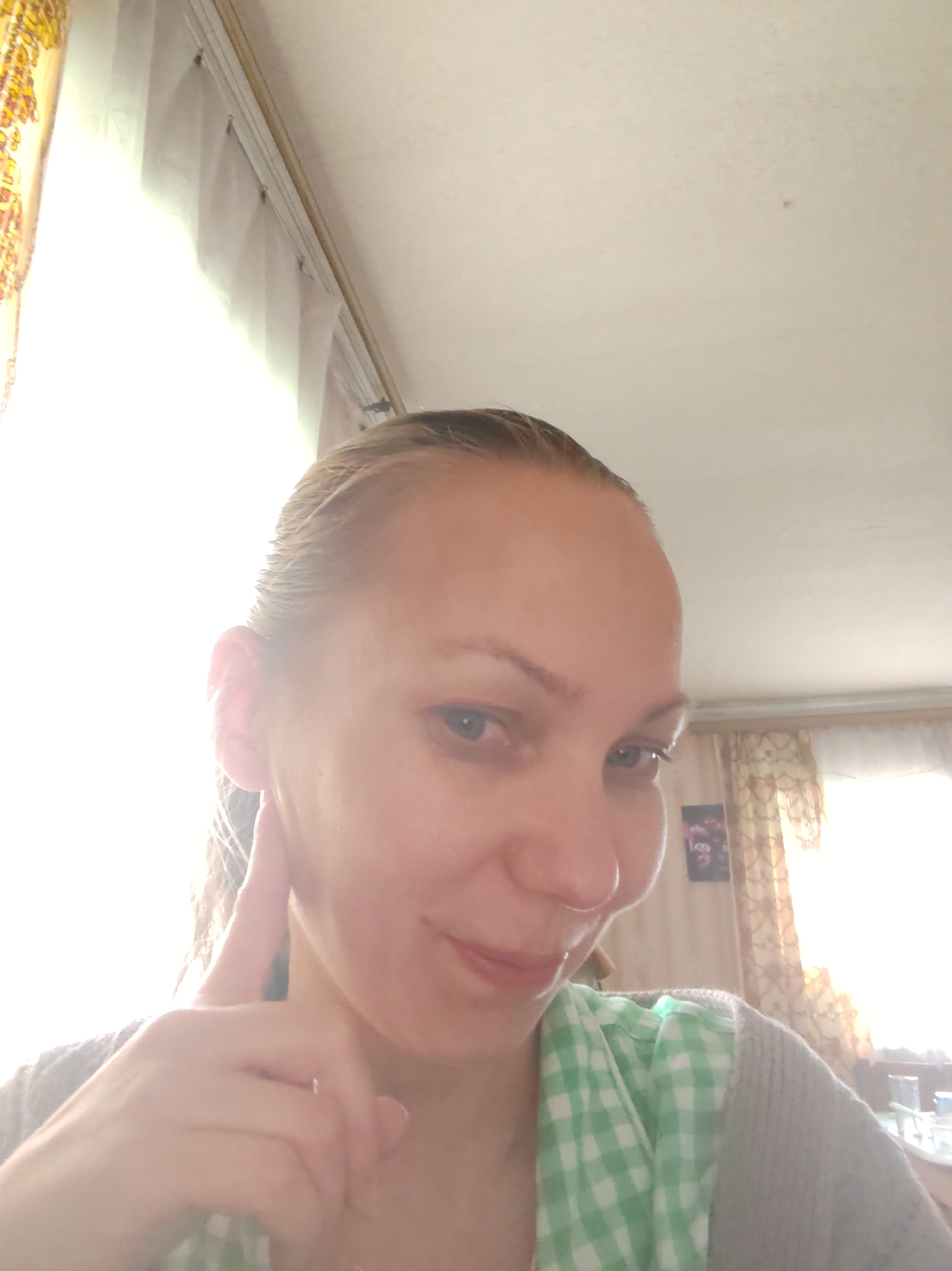 A woman with light skin, light eyes, and blonde hair pulled back is facing the camera. She is touching her right ear with her right index finger. She is wearing a green and white checkered shirt with a grey sweater. The background shows a room with curtains, a window with bright light shining through, and a picture on the wall. Based on the context provided, the location is in Чухлэм, Russia.