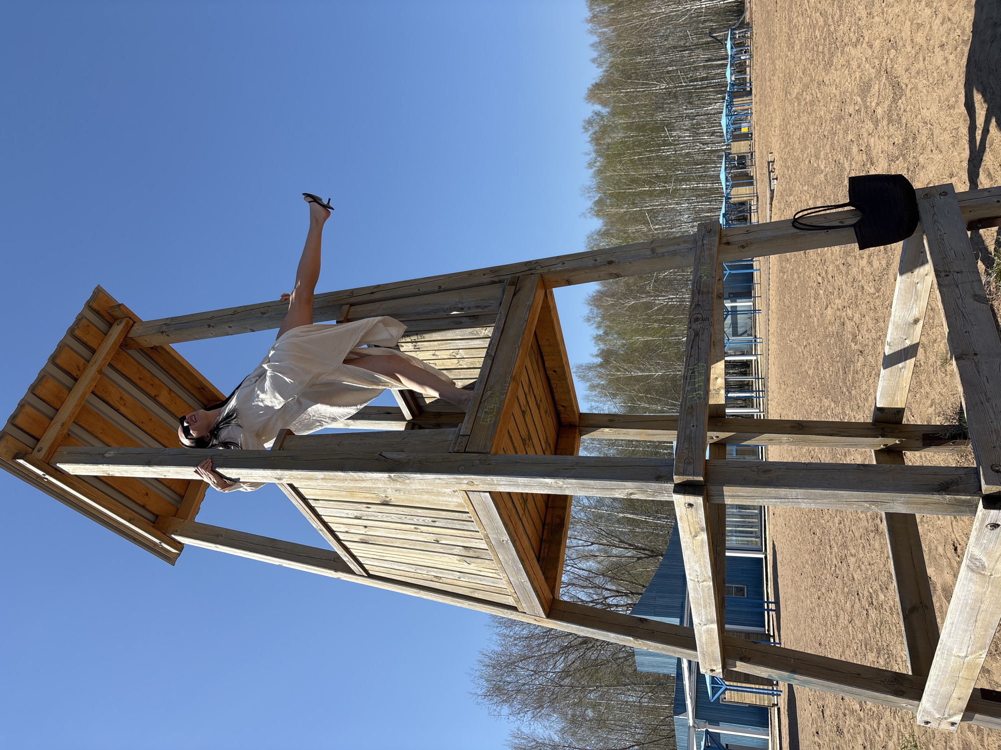 A person is upside down on a wooden structure on a sunny day in Chaikovsky, Russia. The person has one leg extended up in the air, with a black shoe on their foot, and the rest of their body is on the structure. They are wearing a light-colored dress and dark sunglasses. The structure appears to be a playground or lookout tower. There is a black bag hanging on a part of the structure. In the background, there is a sandy area with rows of blue beach shelters, a line of trees, and blue buildings. The sky is clear blue.