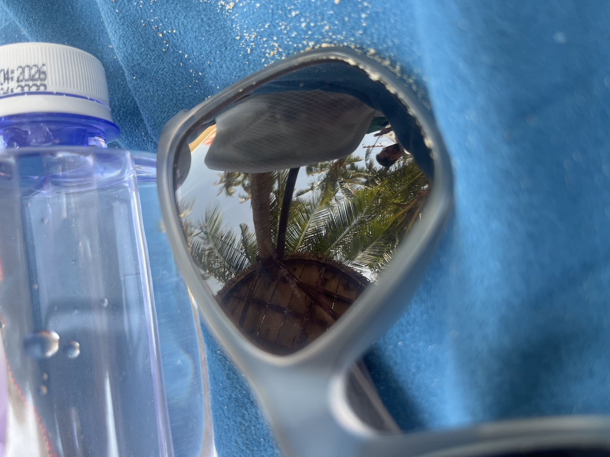 The image shows a pair of sunglasses reflecting a scene of palm trees and a thatched-roof structure. The glasses are lying on a blue towel, next to a bottle of water with a white and blue cap. The date "04:2026" is visible on the water bottle's cap. The reflected scene suggests a tropical location, potentially in Bac Natural Heritage Area Trang, Vietnam, based on the request.