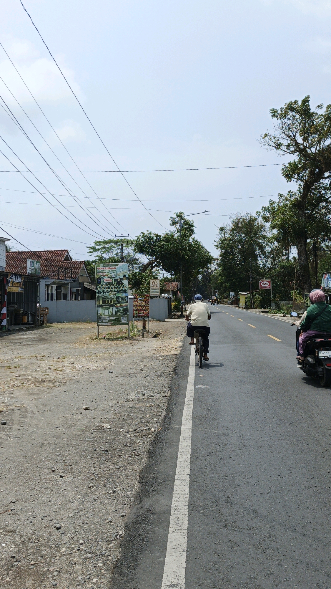 This is a vertical, eye-level shot of a rural road in Sukaresik, Indonesia. The road is paved with asphalt and has a white dashed line marking the lane. The sky is bright and overcast. On the left side of the road, there is a dirt shoulder lined with gravel and rocks. Behind the shoulder, there are some low buildings with red-tiled roofs. A large sign with a group of people in uniform and images of buildings is prominently displayed. Further down the road, on the right, a person on a bicycle is moving away from the camera. Further down the road, another person on a motorcycle is also moving away from the camera. The road is lined with trees on both sides, with some power lines overhead. The overall scene depicts a typical day on a less-traveled road in a village setting.