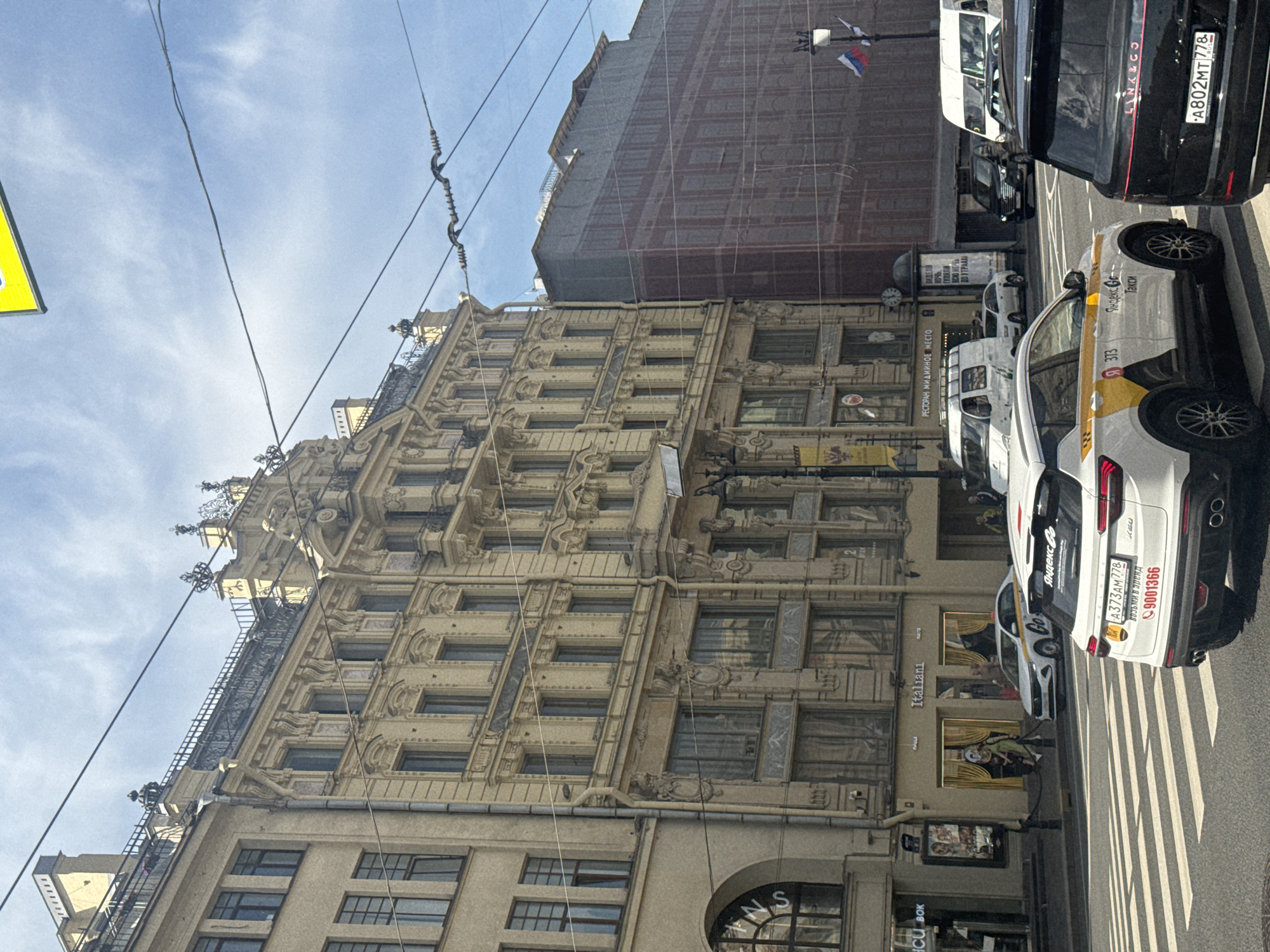 The image depicts a street scene in Saint Petersburg, Russia, on a clear day with a partly cloudy sky. In the foreground, a white Geely Coolray taxi with "Yandex Go" livery is stopped at a pedestrian crossing. The car's license plate reads "A373AM778" and "9001366". To its right, the rear of a black Land Rover is visible, with a license plate "A802MT778". Further down the street, a white Volkswagen Transporter van, a white sedan, and a black SUV are also stopped.
The background features a multi-story building with ornate architecture, typical of Saint Petersburg. The building has many windows and decorative elements. To the right of this building, another structure is covered in scaffolding and a reddish-brown protective mesh, suggesting ongoing renovation or construction. Russian flags are visible on a lamppost in front of the construction site. Signs in Russian are present on the buildings, including one that reads "Italiani" above a storefront. The overall impression is a bustling urban environment with a mix of historic and modern elements.