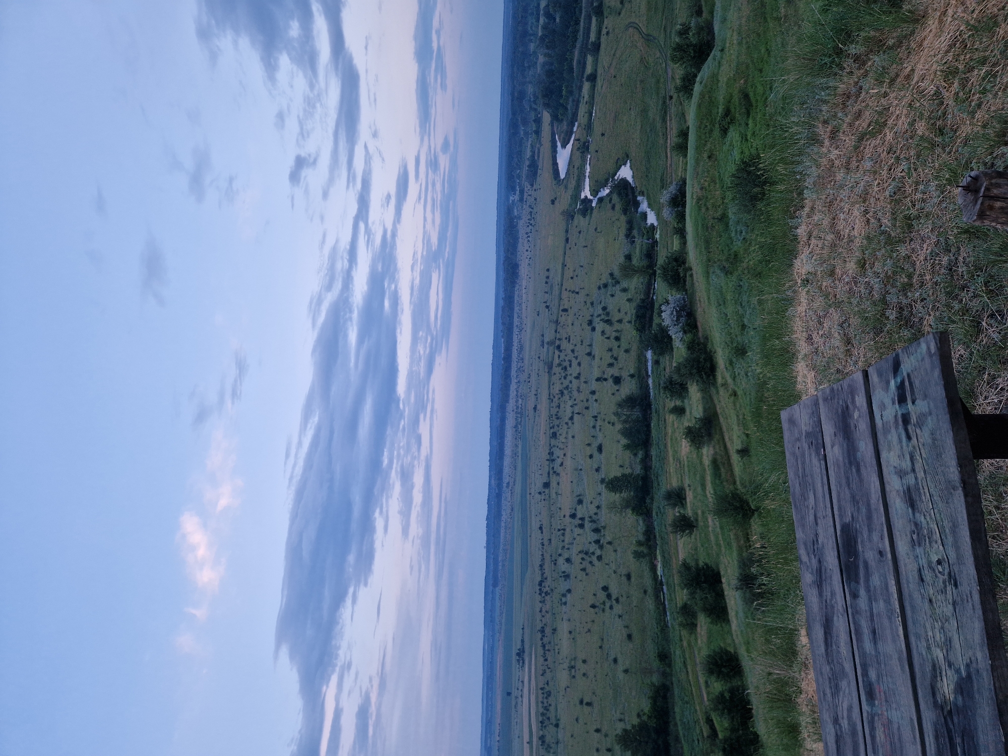 An overhead shot captures a wooden picnic table in the foreground, situated on a grassy knoll. Beyond the table, a sprawling landscape unfolds under a pale blue sky dotted with wispy clouds. A winding river snakes through a verdant plain dotted with scattered trees and vegetation. The scene is bathed in the soft light of dawn or dusk.