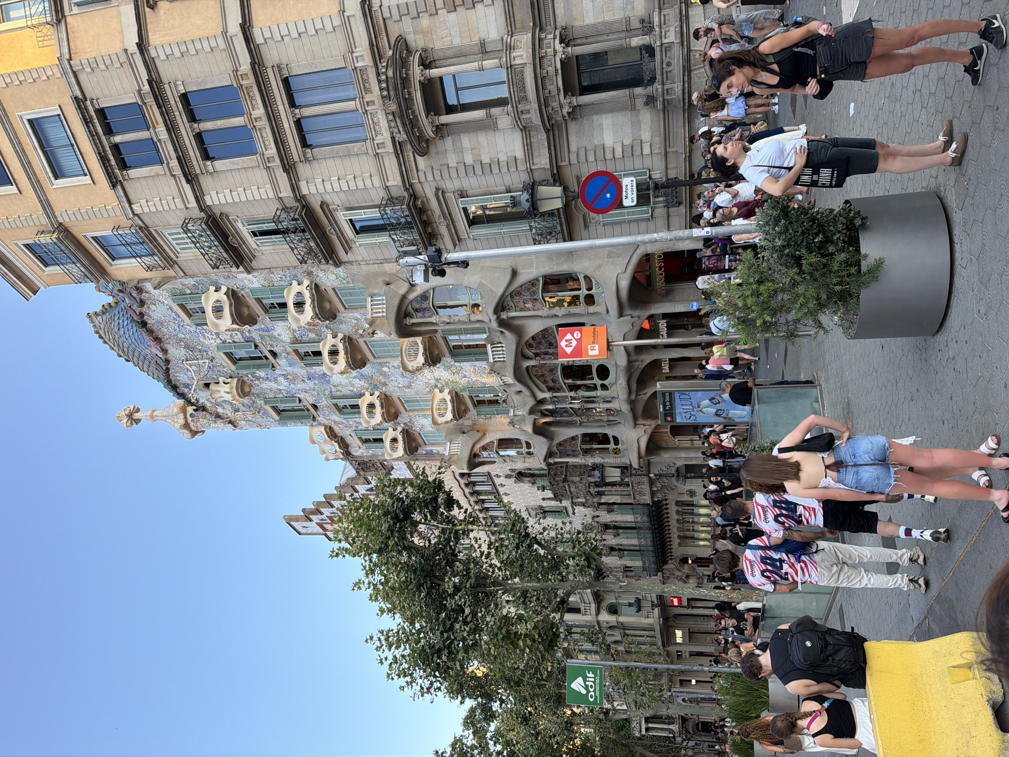 The image captures the exterior of Casa Batlló, a renowned Modernist building designed by Antoni Gaudí, located on Passeig de Gràcia in Barcelona, Spain. The iconic facade, characterized by its organic shapes, colorful mosaic tiles, and skeletal-like balconies, is prominently featured. A street scene unfolds in front of the building, with a clear sky overhead.
Several people are visible on the sidewalk, some walking, others standing and conversing. A group of young adults, including two men wearing matching white t-shirts with red and blue stripes and the number "24," are a focal point. One woman is seen from behind, wearing denim shorts and a white top. Other individuals are scattered throughout the frame, dressed in casual summer attire.
Street furniture and signage are present, including a green "adif" sign on a pole, a red and orange metro sign, and a circular "no motorcycles" sign. Large planters with green foliage are also part of the urban landscape. The building next to Casa Batlló exhibits a more conventional architectural style, with a beige facade and symmetrical windows. The overall scene depicts a bustling urban environment with significant architectural interest.