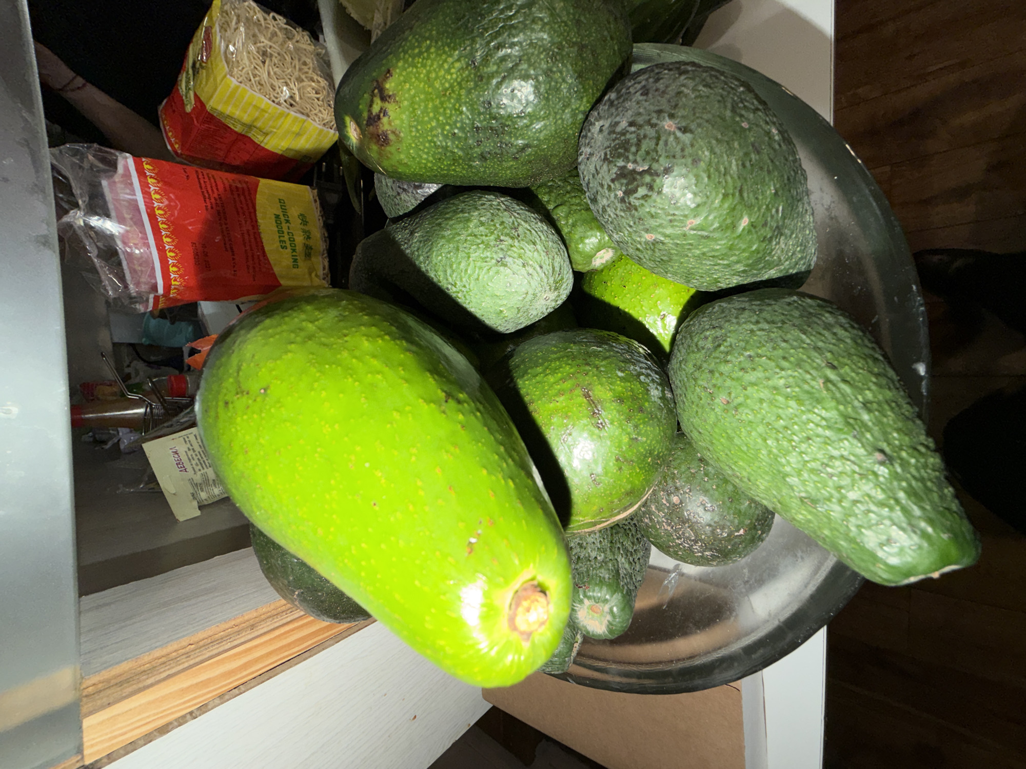 A tilted, high-angle shot reveals a stainless steel bowl brimming with avocados of varying shades of green and ripeness, ranging from a bright, almost yellow-green to a darker, mottled green. The bowl rests on a white surface, likely a countertop or cabinet. Beside the bowl, to the left, are packaged noodles with red and yellow packaging. Other items are visible in the background, which appears to be a storage area or cupboard. The location and city information is irrelevant to the visual content of the image.