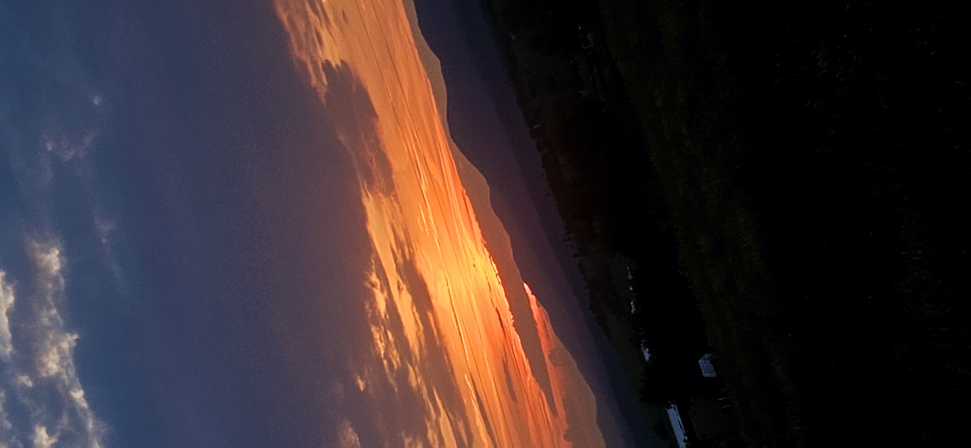 A wide-angle landscape photograph, taken at sunset, depicts a mountainous horizon under a vibrant sky. The upper left quadrant of the sky is dominated by dark blue and purple hues with scattered, illuminated cumulus clouds, hinting at an ongoing transition from day to night. Towards the center and right, the sky transitions into an intense orange and red palette, indicative of a sunset, with elongated, horizontal cloud formations absorbing and reflecting the light. The sun's position is obscured by a distant mountain range, creating a bright orange glow just above the silhouette of the peaks.
The mid-ground features multiple layers of mountains, presented in progressively darker shades of deep purple and blue-black as they recede into the distance, demonstrating atmospheric perspective. The most distant mountains are sharply silhouetted against the bright horizon.
The foreground, occupying approximately the lower third of the image, is heavily shadowed, appearing almost entirely black. Despite the low light, faint outlines of dark green vegetation, likely grass or low shrubs, are discernible. A few indistinct, rectangular structures with light-colored roofs, possibly buildings or trailers, are visible in the far left of the shadowed mid-ground, nestled among darker vegetation. No people or animals are visible. The scene captures a tranquil, natural landscape at dusk, in the vicinity of Momin Prohod, Bulgaria.