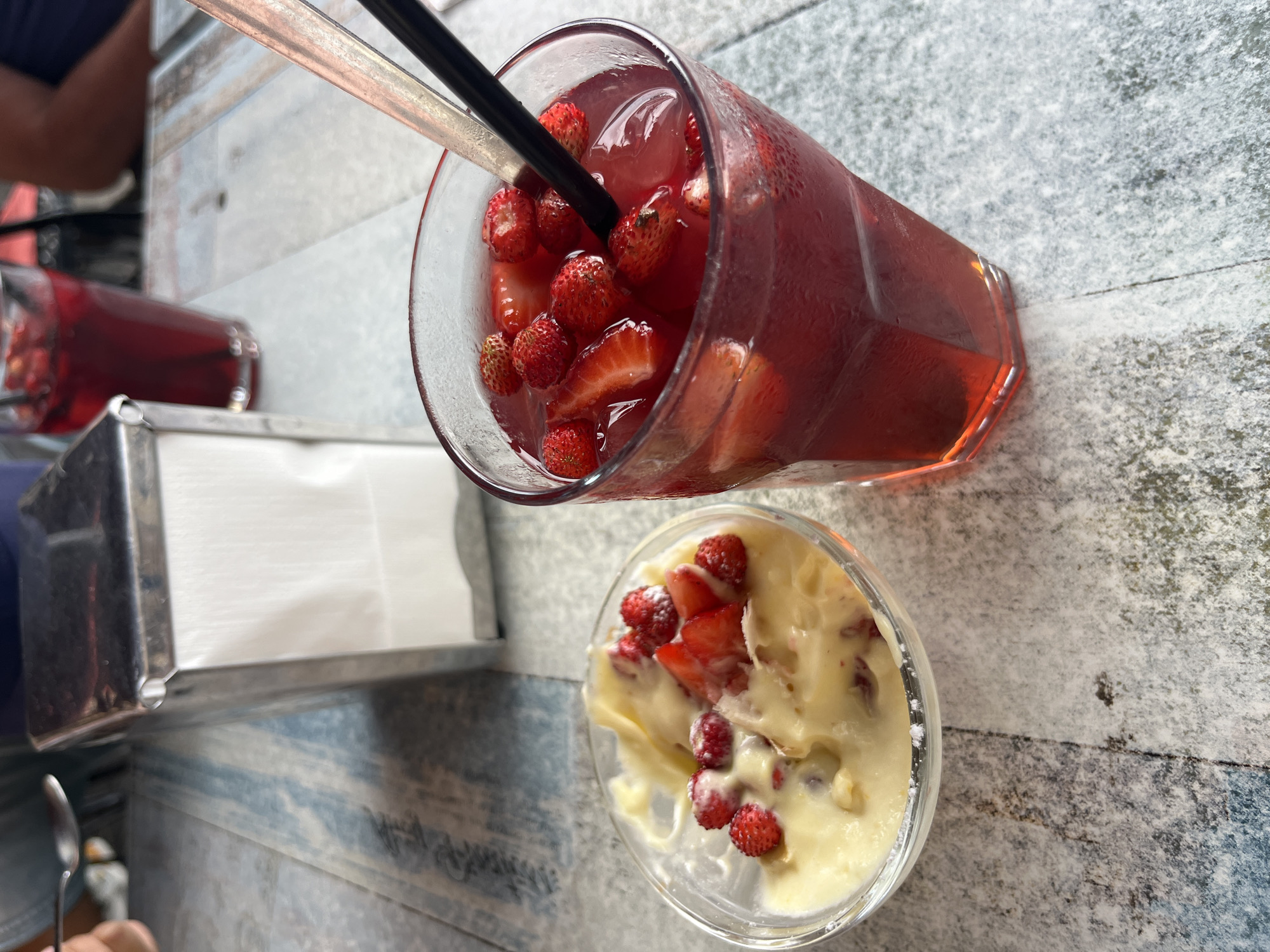 On a table with a distressed blue and white patterned surface, two drinks and a dessert are arranged. To the left is a small glass bowl containing a light-colored, creamy dessert topped with sliced strawberries and whole wild strawberries. Next to it, to the right, is a tall glass filled with a red beverage, likely a drink with strawberries and ice. Two straws, one black and one silver, protrude from the glass. In the background, slightly out of focus, is a stainless steel napkin dispenser. The overall impression is of a refreshing treat enjoyed outdoors, possibly at a cafe in Nemi, Italy, known for its strawberries and proximity to Lake Nemi. The lighting suggests it is daytime.