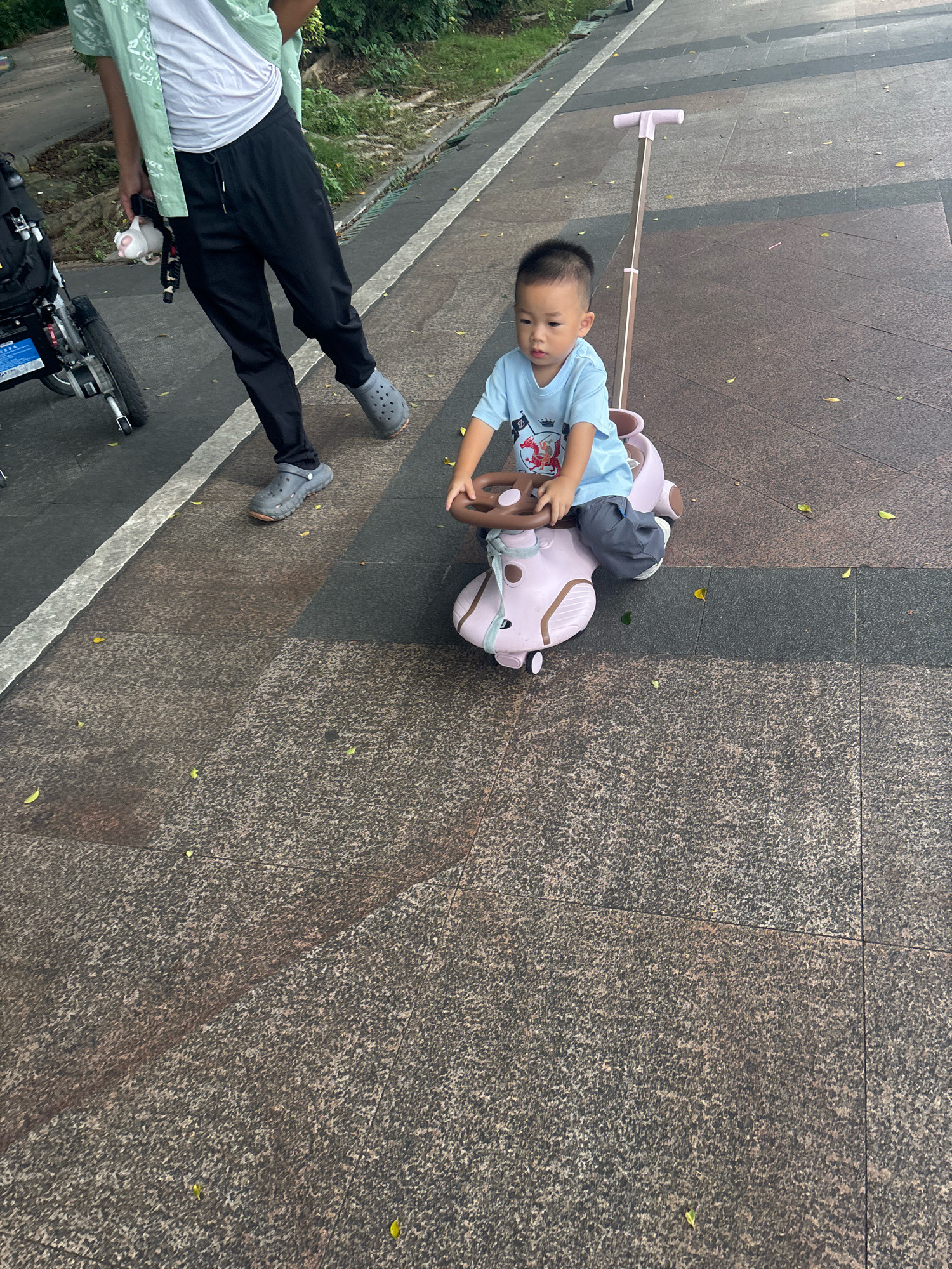 An outdoor, ground-level, slightly high-angle shot captured on a paved walkway in Nanhai District, China, shows a young boy riding a pink toy car. The boy, with short dark hair and wearing a light blue t-shirt and gray pants, is seated in the toy car, his hands on the steering wheel. The toy car has a pink handle extending upwards behind it. To the left of the frame, an adult, partially visible, is walking alongside a black stroller. The adult is wearing a green patterned shirt, white t-shirt, and black pants, and gray Crocs. The ground is paved with textured, dark gray and brown stone tiles, with scattered fallen leaves. A white line marks the edge of the walkway. The overall lighting suggests a moderately sunny day.