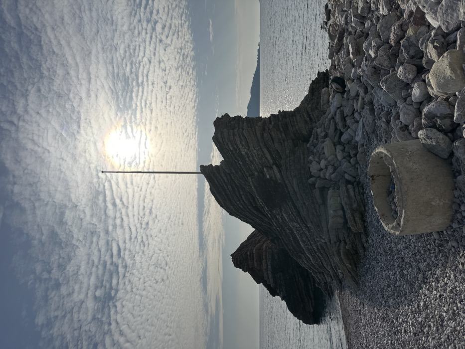 The image depicts a coastal scene in Bečići, Montenegro, under a partially cloudy sky. The sun, visible through a thin cloud layer, forms a bright halo and casts a strong reflection across the calm surface of the Adriatic Sea to the right.
In the foreground and extending to the left, a pebble beach is visible, composed of irregularly shaped light and dark grey stones. A weathered, cylindrical concrete ring, possibly a culvert section, rests on the pebbles in the immediate foreground, containing some indeterminate debris.
Dominating the midground are two large, dark rock formations with distinct horizontal geological stratification. The larger formation, centrally located, is topped by a tall, slender, dark-colored pole or mast extending vertically towards the sky. The second, slightly smaller rock formation is to the left, partially submerged in the seawater.
In the distant background, across the sea to the right, a hazy, mountainous landmass with visible structures along its base is present on the horizon. No individuals, animals, or active events are depicted in the scene.