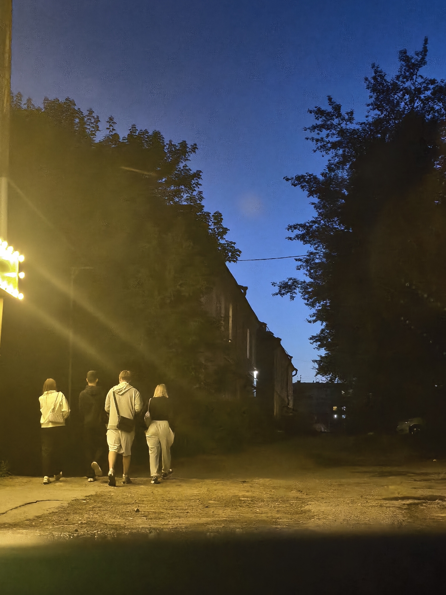 A group of four individuals, two male and two female, walk away from the camera on a dirt path at dusk in an urban environment. The individuals are dressed in casual clothing. A string of lights is visible to the left of the frame, casting a bright yellow glow on the scene. Behind the individuals, a brick building is visible, with a large tree to its right, and further buildings in the background. The sky is a deep blue, indicating it is twilight.