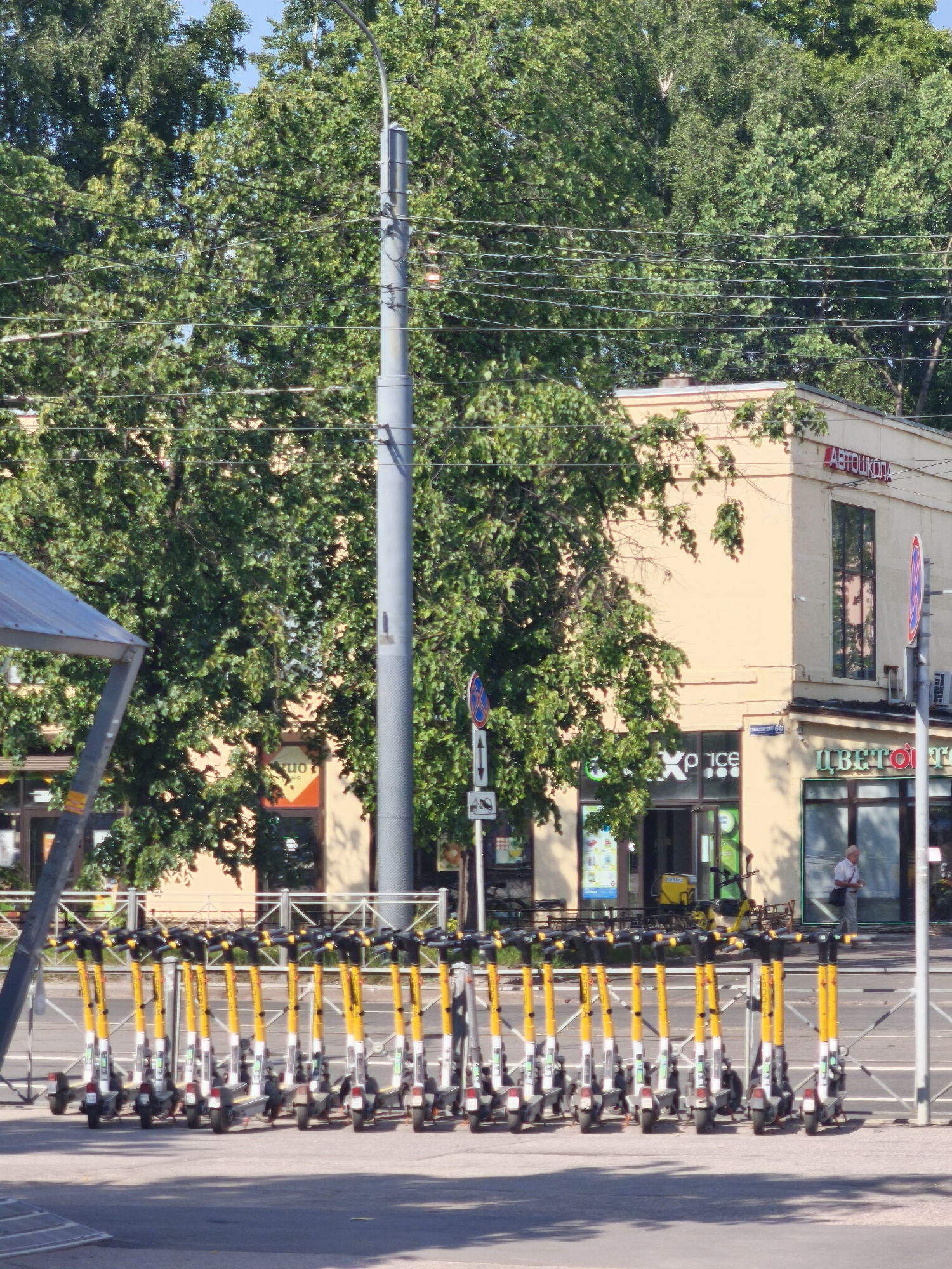 The image shows a row of yellow and white electric scooters parked on a sidewalk in front of a light yellow building. The building has storefronts with signs that read "ABTOIKON", "max price", and "ЦВЕТО". A person in a light-colored shirt and pants is walking in front of the "ЦВЕТО" store. There are trees in the background and a lamppost on the left side of the image. The scene appears to be a street in Saint Petersburg, Russia.