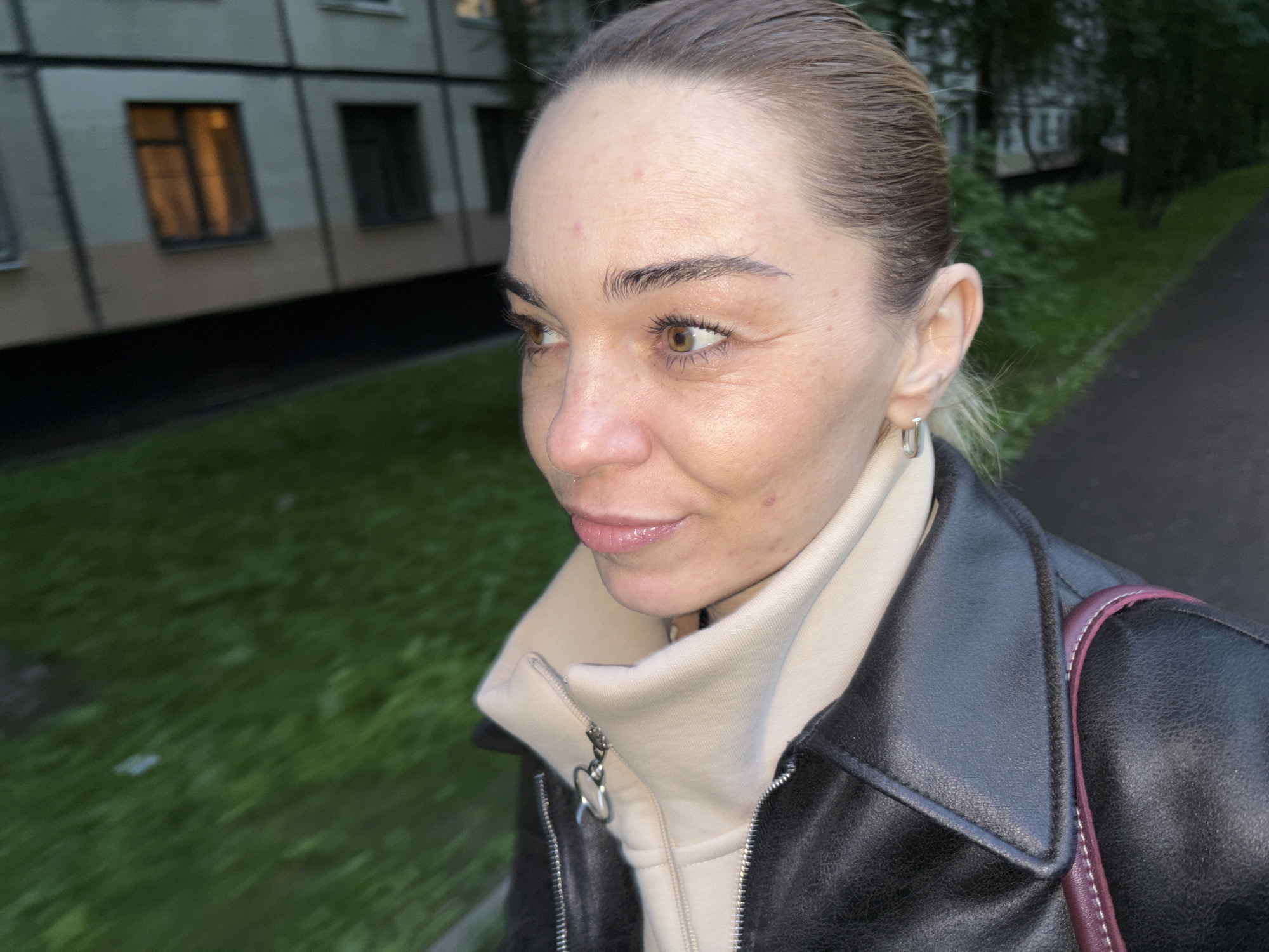 A medium shot captures a woman in an outdoor urban setting. She is turned to her right, looking slightly upwards, her expression relaxed and her lips parted. Her hair is pulled back, and she wears a dark leather jacket over a light-colored hooded garment. Her left ear is adorned with a small silver hoop earring. In the background, a building with multiple windows is visible, with one window emitting a warm, orange light. To the right of the building, a green, grassy area lines a paved path, suggesting a park or sidewalk. The overall lighting is dim, indicating it may be evening or overcast.