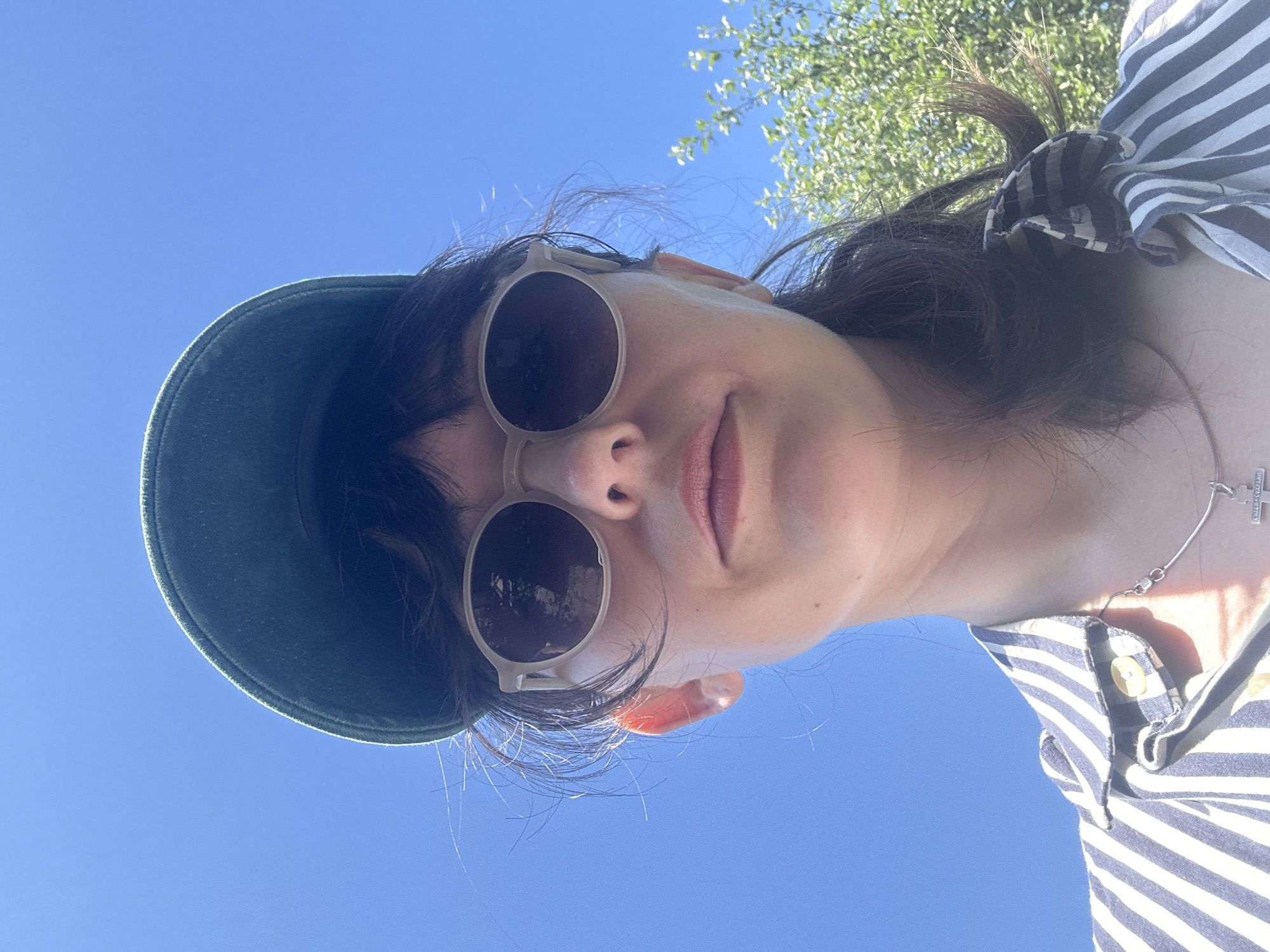 This is a close-up, low-angle selfie of a young woman outdoors, likely on a sunny day. She is wearing a dark green baseball cap and light-colored round sunglasses. Her hair is dark and appears somewhat windswept. She is also wearing a striped shirt with buttons and a necklace with a cross pendant. The background consists of a clear blue sky and the green foliage of a tree. The woman's expression is neutral, with her lips slightly parted. The image is shot from a bottom-up perspective, with the sky taking up a significant portion of the frame. There is no visible text in the image.