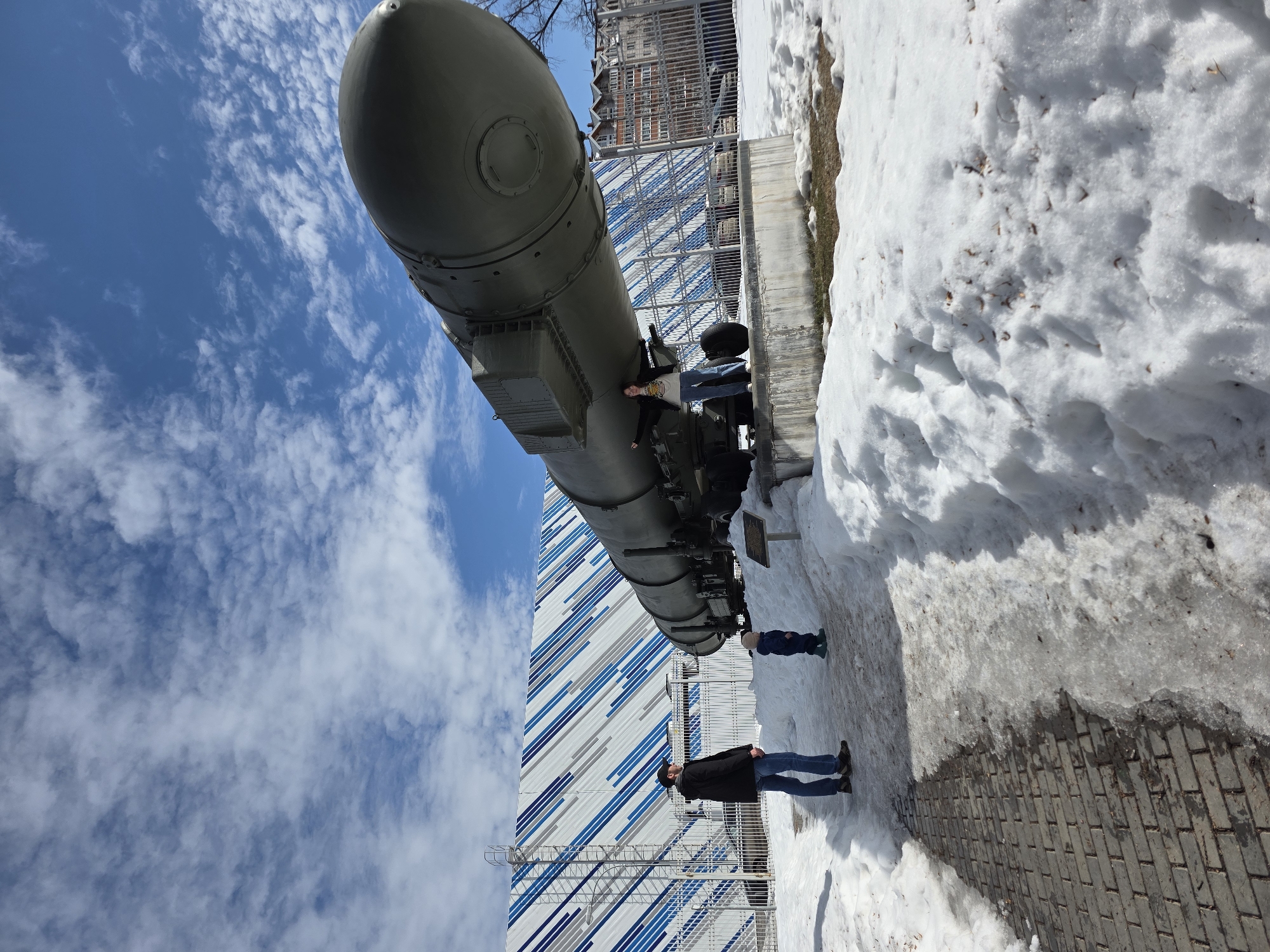 The image displays an outdoor scene in Votkinsk, Russia, at what appears to be a public exhibit or memorial associated with the Votkinsk Machine Building Plant. The primary subject is a large, olive-drab cylindrical object, identified as an SS-20 Saber (RSD-10 Pioneer) intermediate-range ballistic missile, or a replica, displayed horizontally on a concrete base with a multi-wheeled transport trailer.
Three individuals are present. On the left, a man wearing a black jacket, dark baseball cap, blue jeans, and brown shoes stands on a paved brick walkway, looking towards the missile. Adjacent to him, a small child in a blue hooded outfit and green boots also observes the missile. On the right, a woman stands on the concrete base next to the missile, with her arms outstretched. She wears a dark jacket, a light-colored t-shirt with a graphic design, blue jeans, and white sneakers.
The ground is partially covered in snow and patches of melting ice, indicating a winter or early spring setting. The sky overhead is clear blue with scattered white cumulus clouds. In the background, behind a metal fence, a large building with a modern facade featuring diagonal blue, white, and grey stripes is visible. To the far right, additional multi-story buildings with red roofs are present. A small informational plaque is positioned on the ground near the missile's base, between the man and the exhibit.